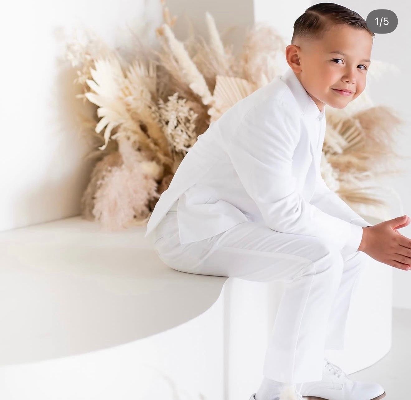 Young boy in a white suit sitting on a white bench with dried pampas grass decor in the background.
