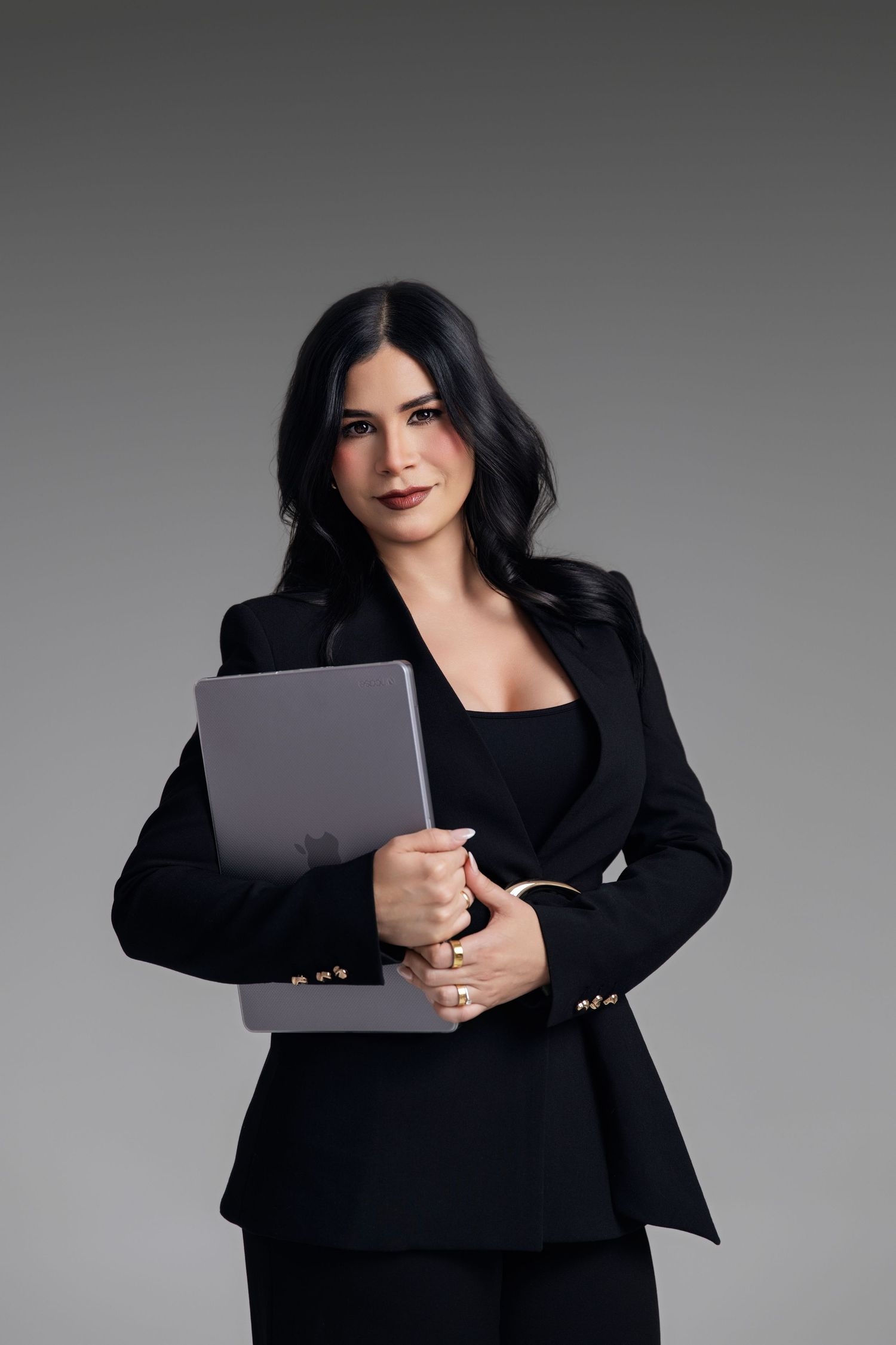Professional woman in black blazer holding a MacBook laptop, posing confidently against a gray background.