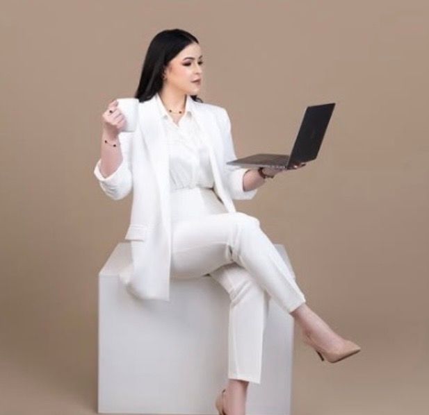 Woman in a white blazer and trousers sits on a white cube holding a coffee cup and laptop against a neutral beige studio backdrop.