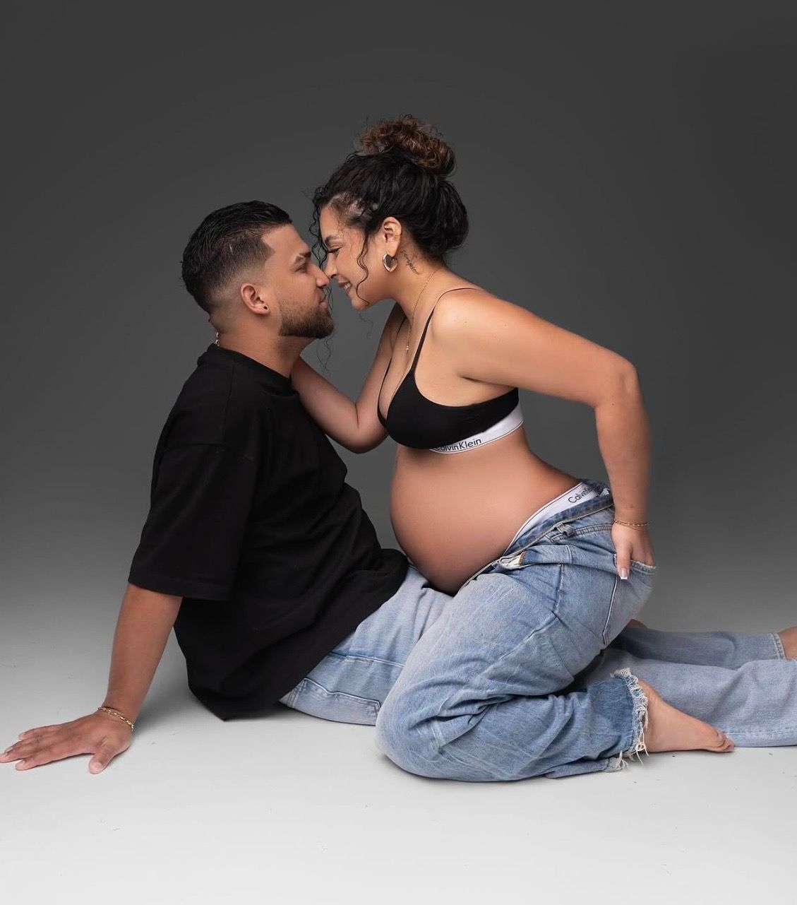 Pregnant woman in a black top and jeans touches noses with her partner during a maternity photoshoot against a gray studio backdrop.