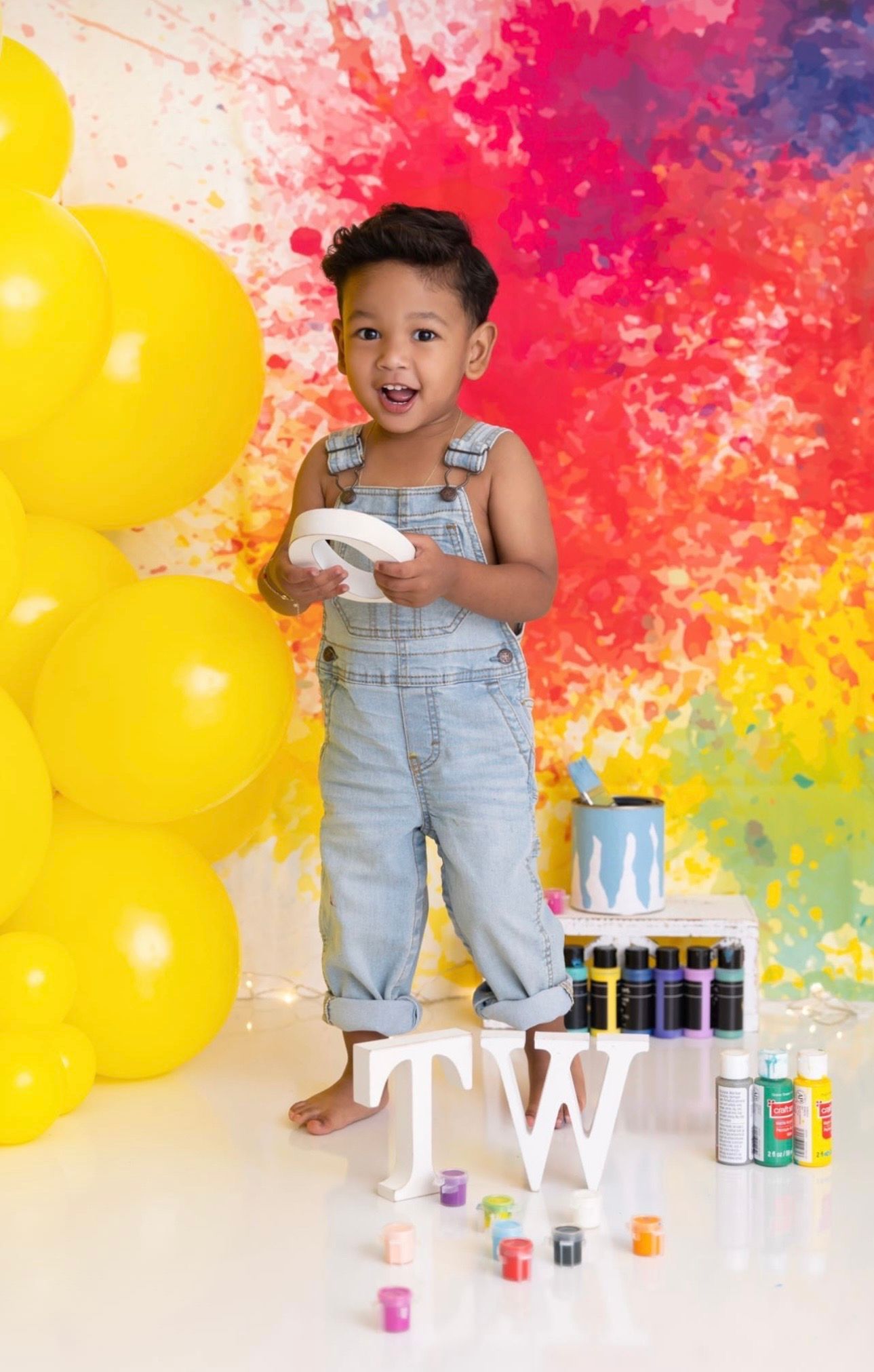 Smiling toddler in denim overalls holds a white letter beside yellow balloons, a colorful paint splash backdrop, paint bottles and letters T