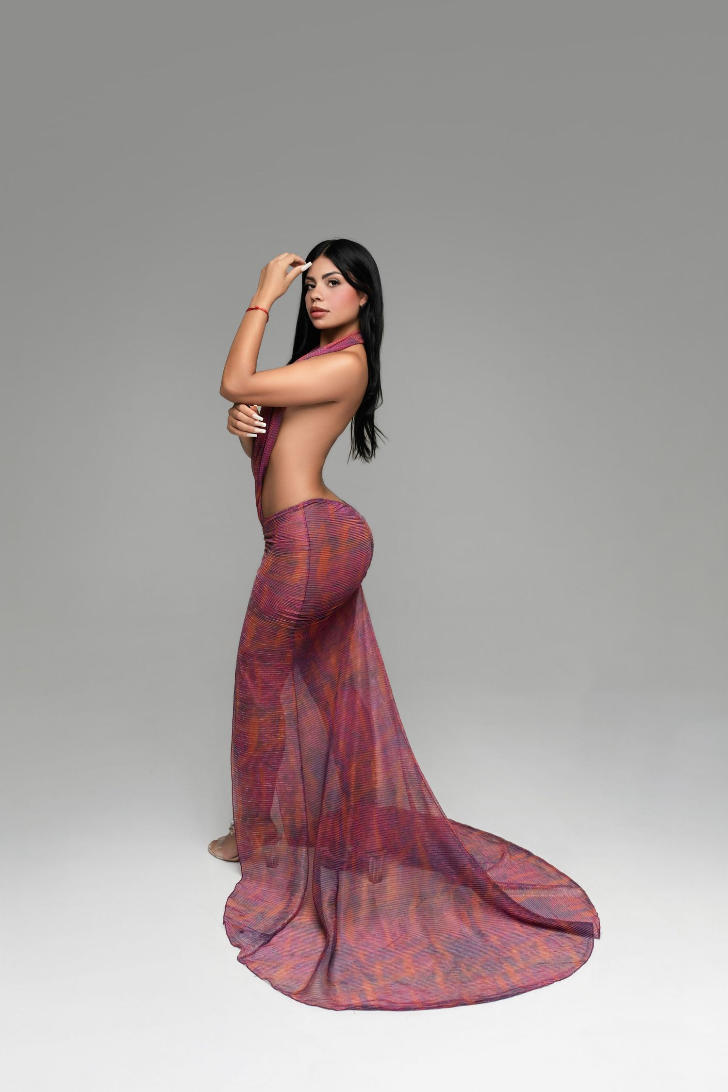 Woman with long dark hair poses in a backless magenta and orange sheer gown with a flowing train against a gray studio backdrop.