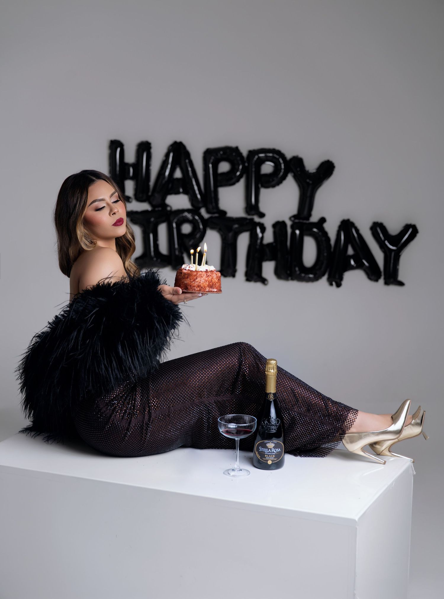 Woman in a black feather-trimmed dress holds a lit birthday cake beside champagne and black Happy Birthday balloons.