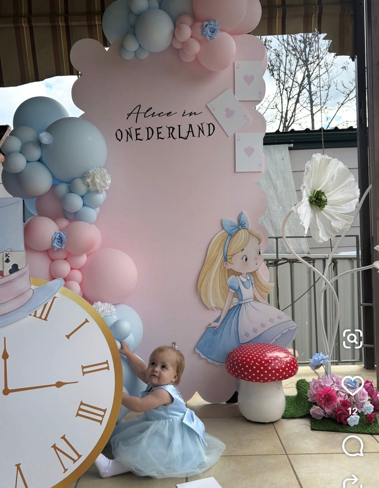 Baby girl in blue dress at Alice in Onederland 1st birthday party with balloon arch and themed decorations.