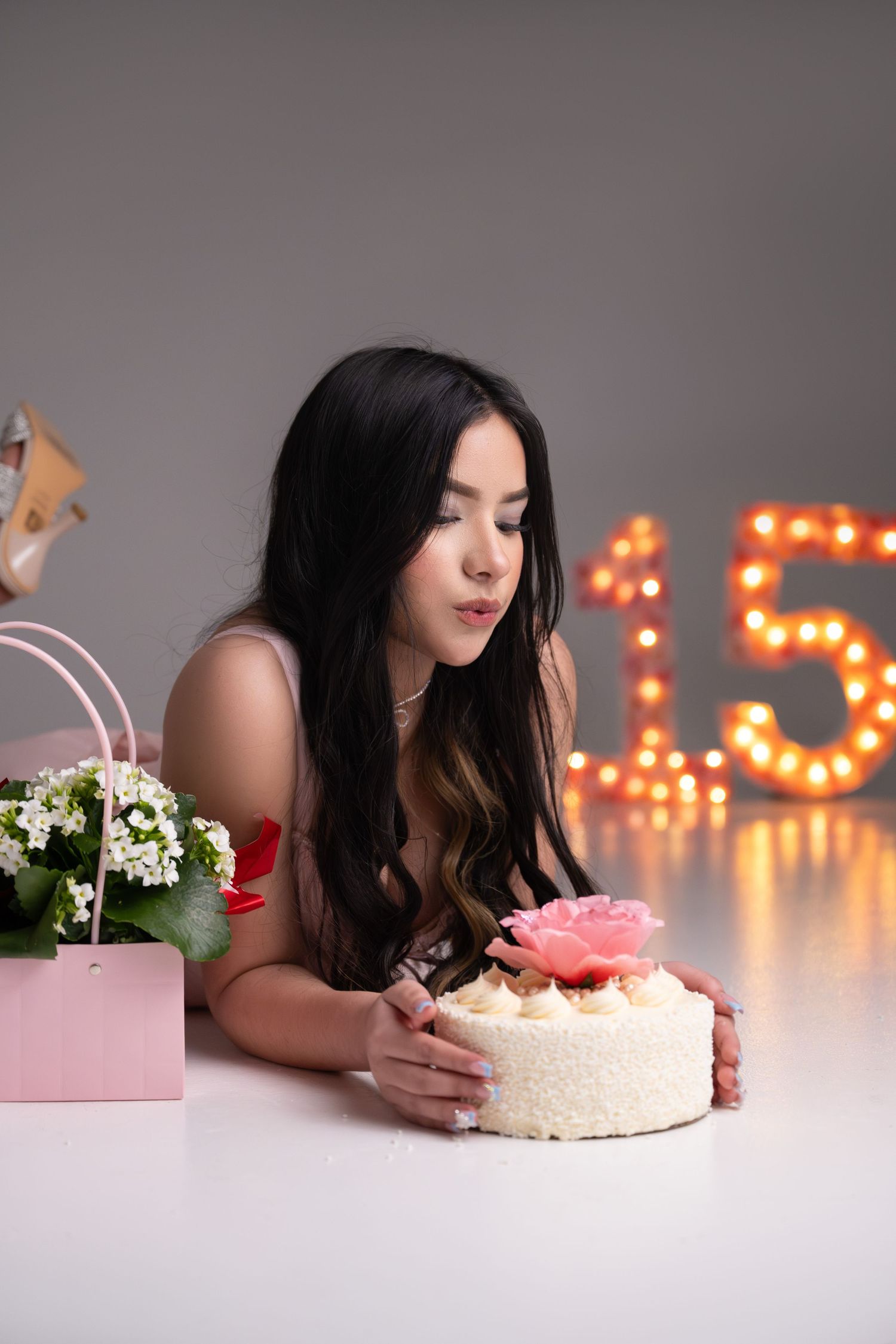 Young woman with long dark hair admires a white floral cake beside flowers and glowing number 15 decorations.