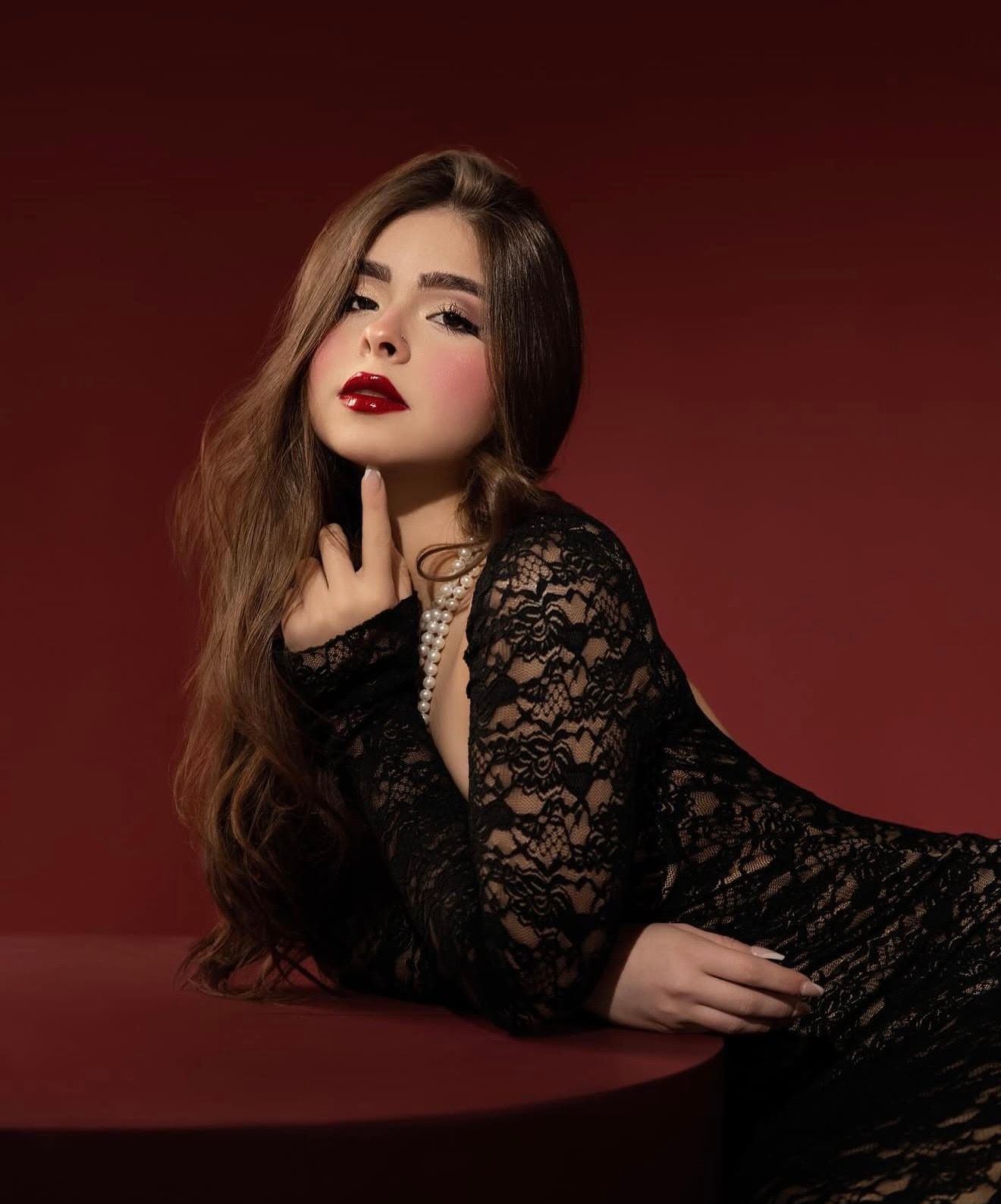 Young woman in black lace dress with pearl necklace poses elegantly against a deep red background with bold makeup.