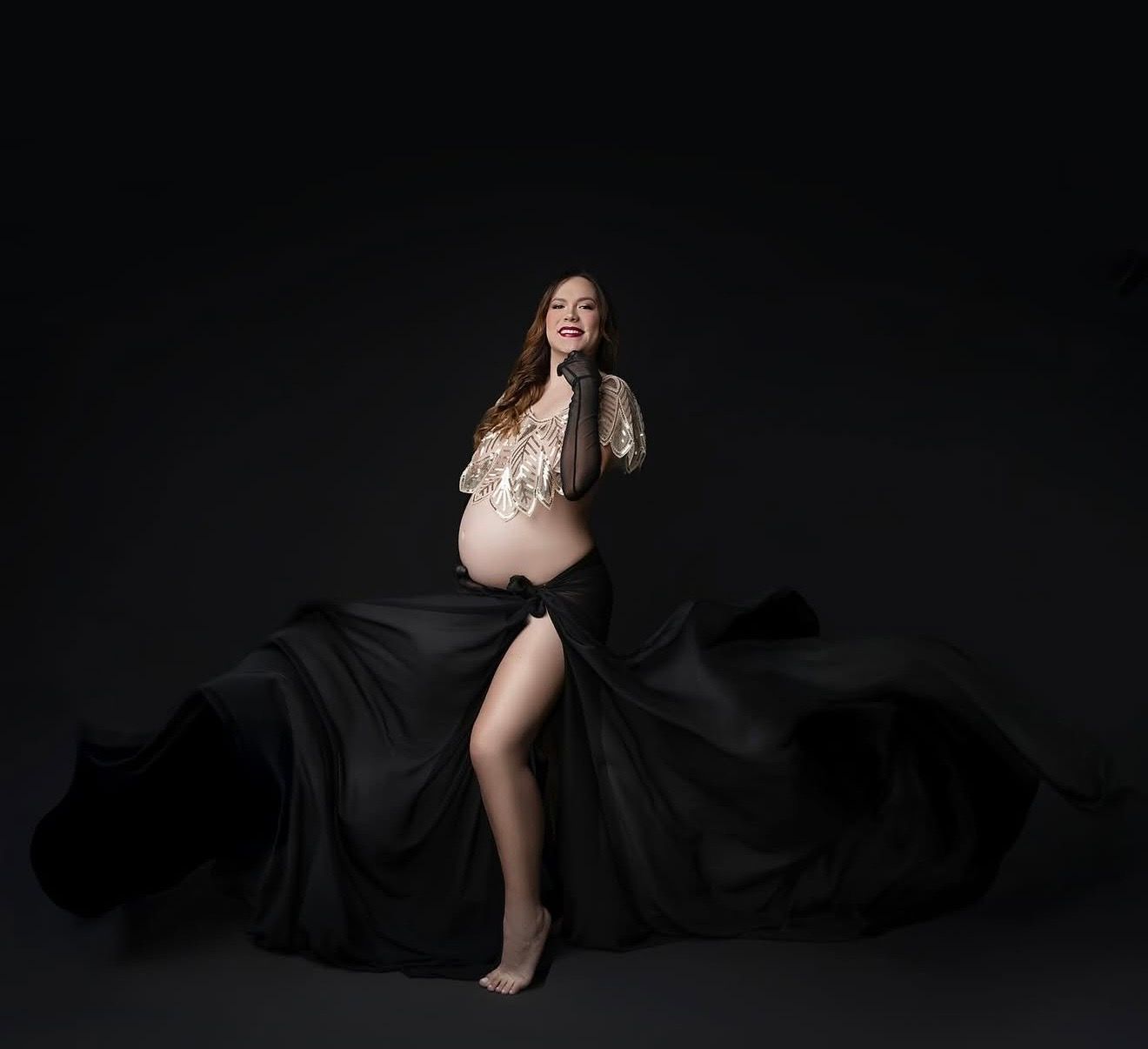 Pregnant woman in black flowing skirt and gold crop top posing gracefully in a professional maternity photo studio.