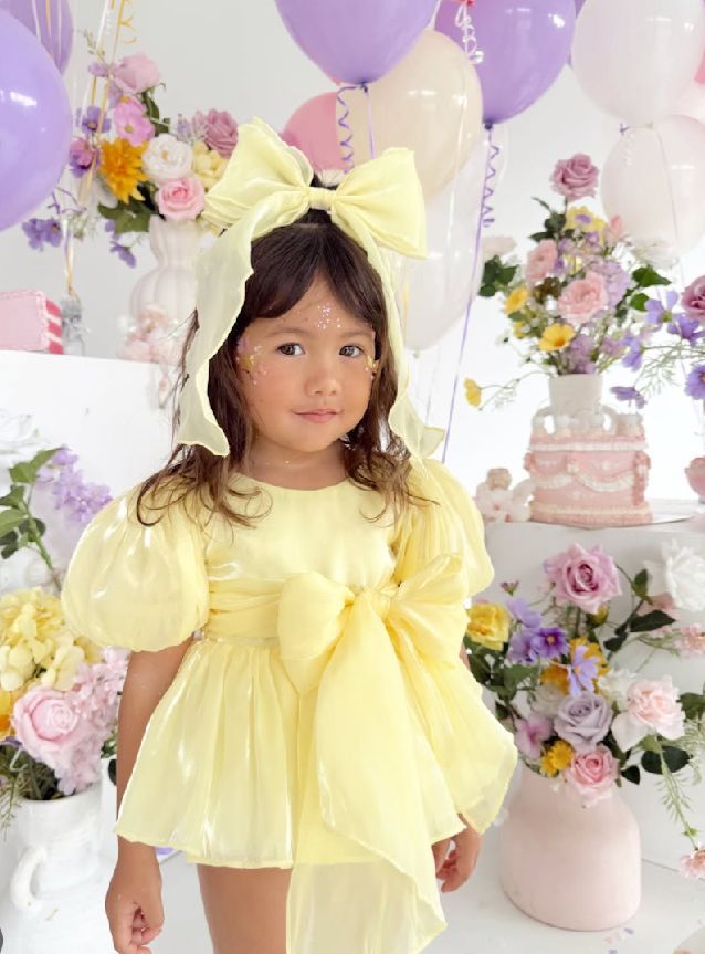 Pastel party setting with yellow dress, purple balloons, and pink floral arrangements in a dreamy backdrop.