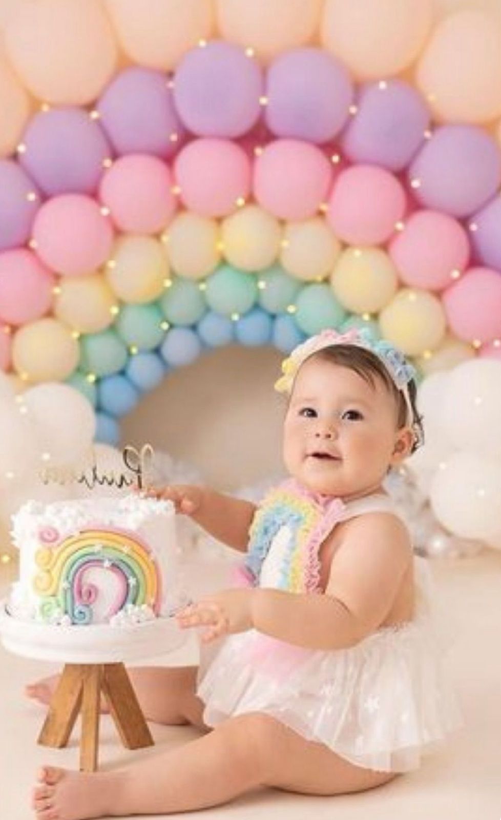 Rainbow balloon arch decorates a whimsical cake smash photo setup with pastel colors and wooden easel.
