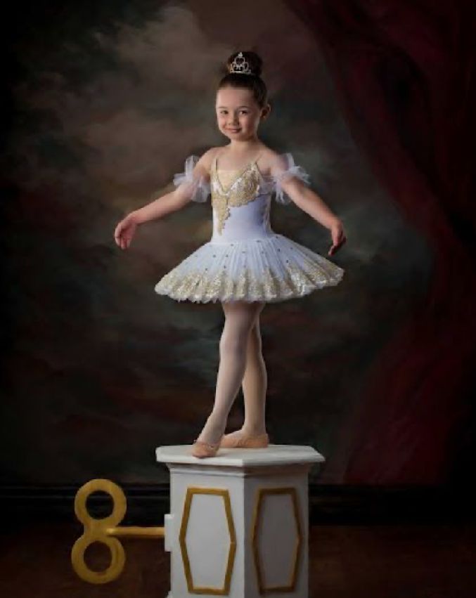A young ballet dancer in a white tutu dress performs graceful poses on a small white stool against a dark backdrop.