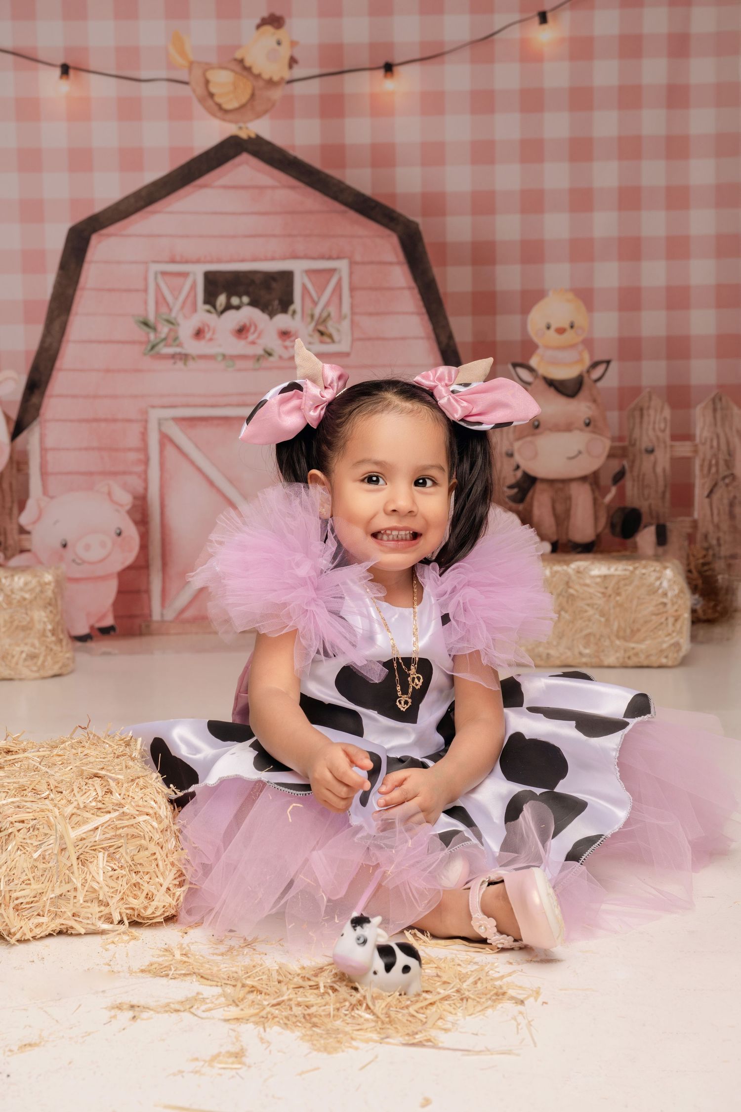Toddler girl in pink tutu cow print dress sits on hay bales in a farm-themed photo shoot backdrop.