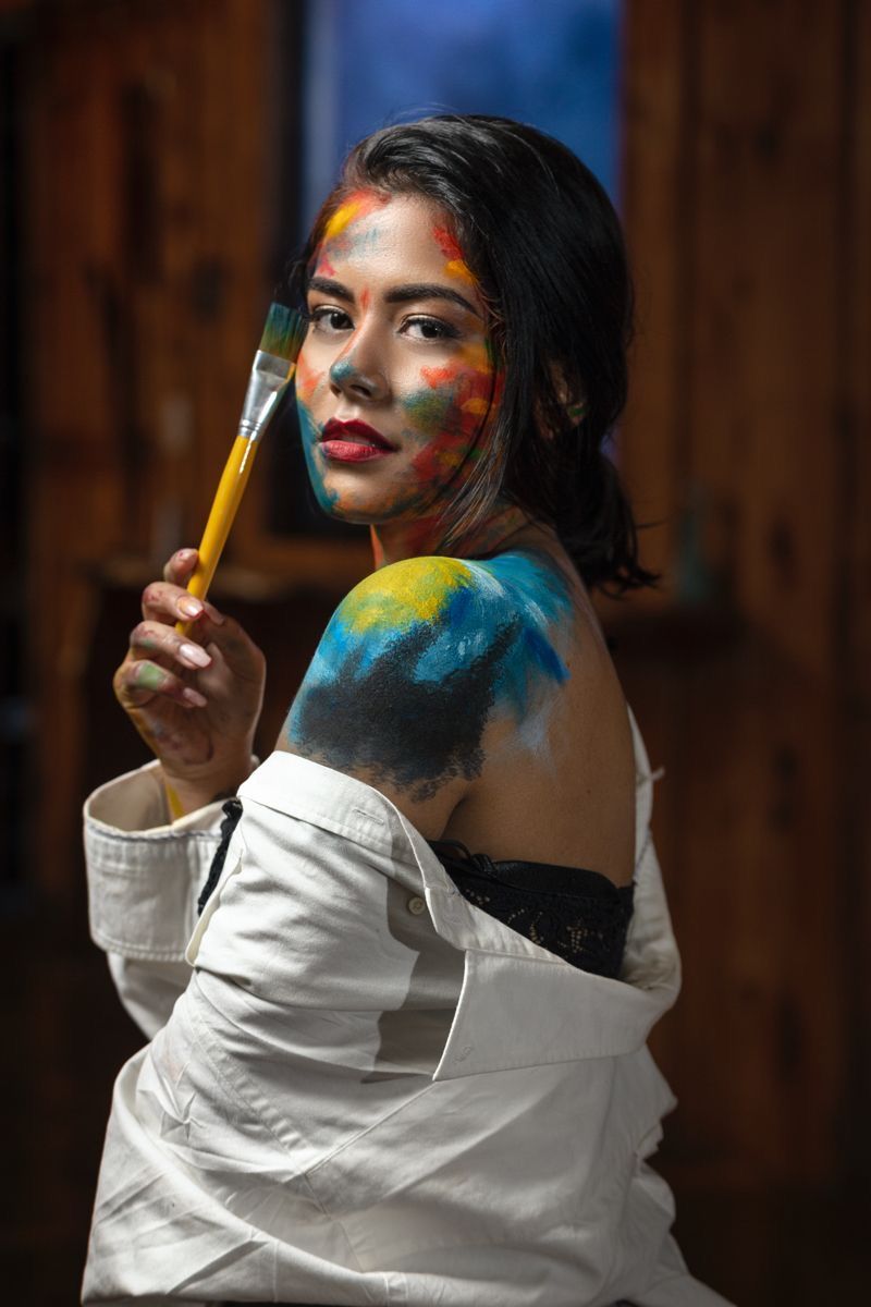 Young woman artist with colorful paint on face and shoulder holding paintbrush in white shirt.