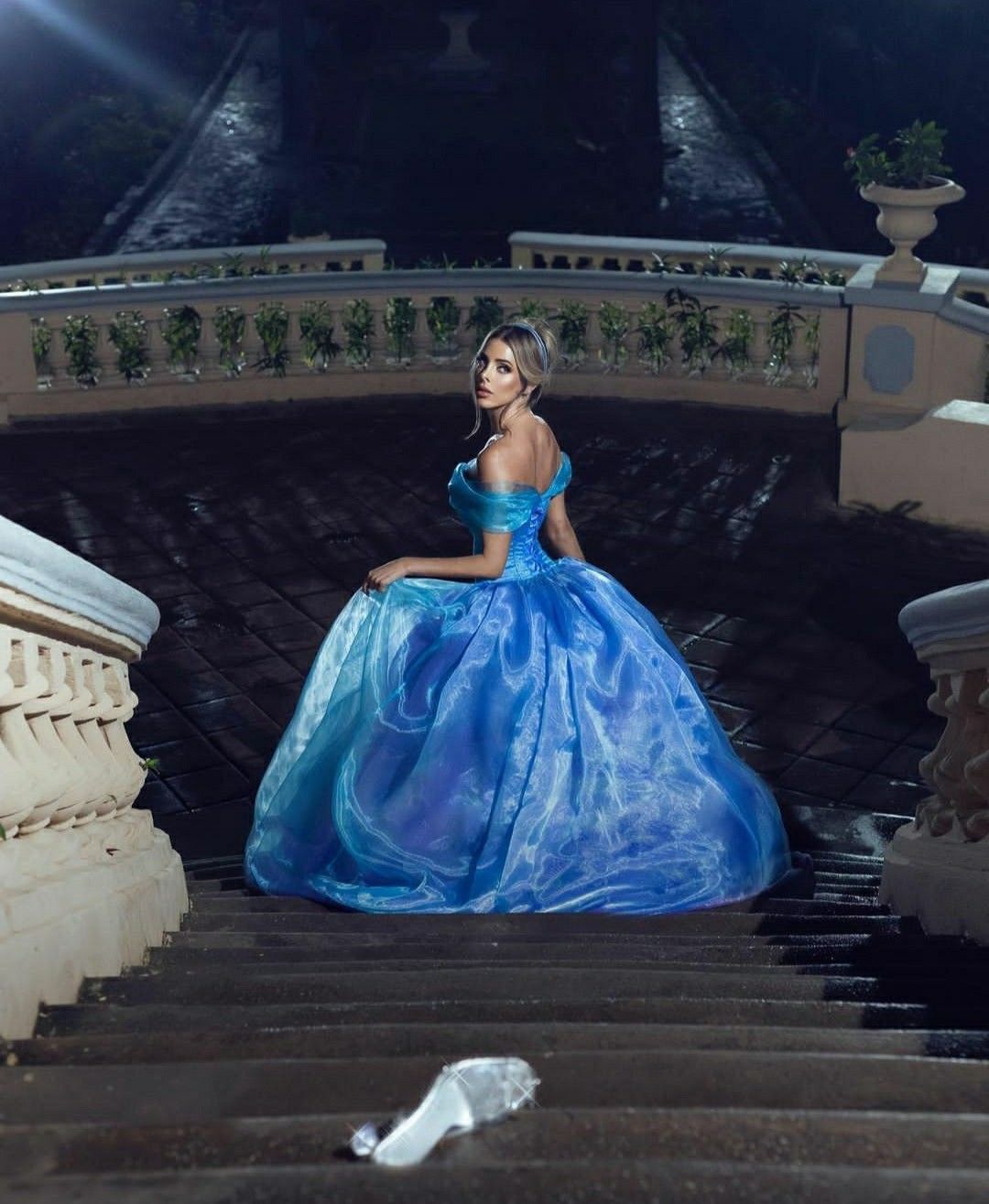 Woman in blue ball gown on grand staircase at night, resembling Cinderella with a glass slipper nearby.
