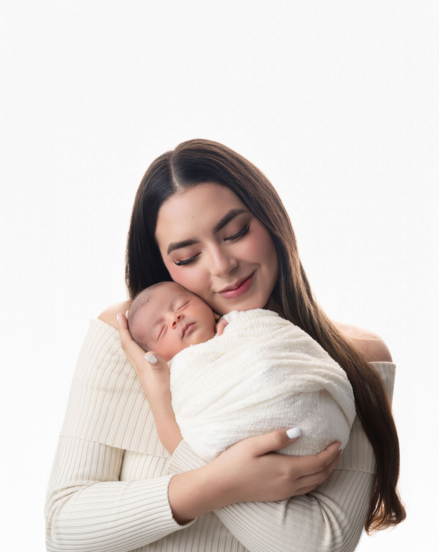 A mother with long dark hair tenderly holds her sleeping newborn wrapped in white against a clean white background.