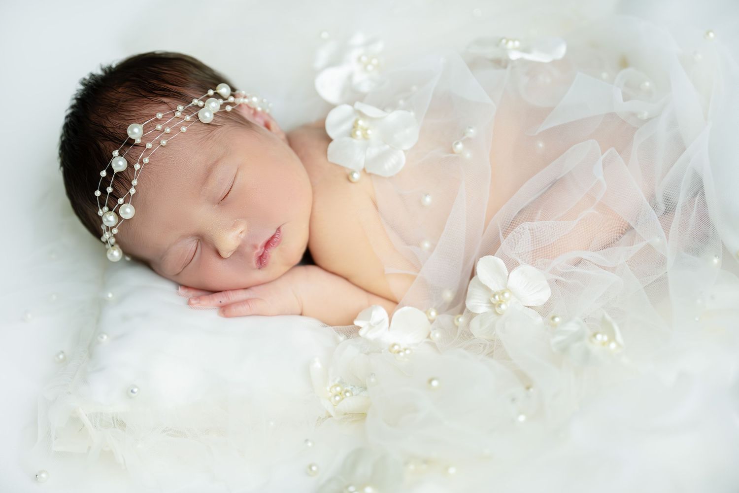 Peaceful newborn baby sleeping on white fur, adorned with a delicate floral pearl headband and white tulle wrap.