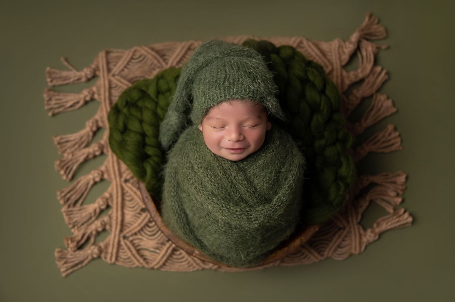 Newborn baby wrapped in green knit swaddle with matching hat, posed on a beige fringe blanket with green wool accents.