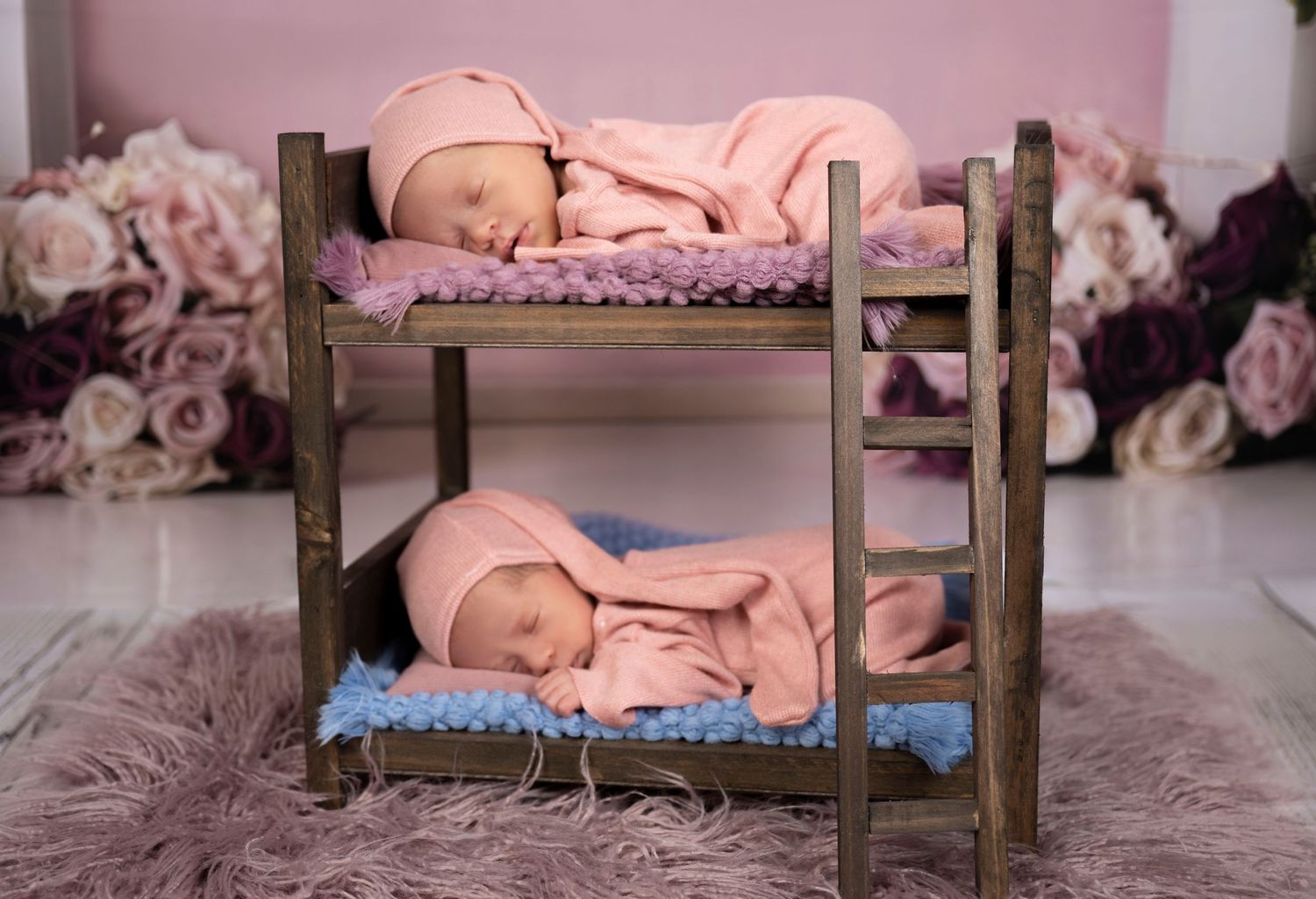 Two newborn twins sleeping peacefully in pink outfits on a miniature wooden bunk bed with floral backdrop.