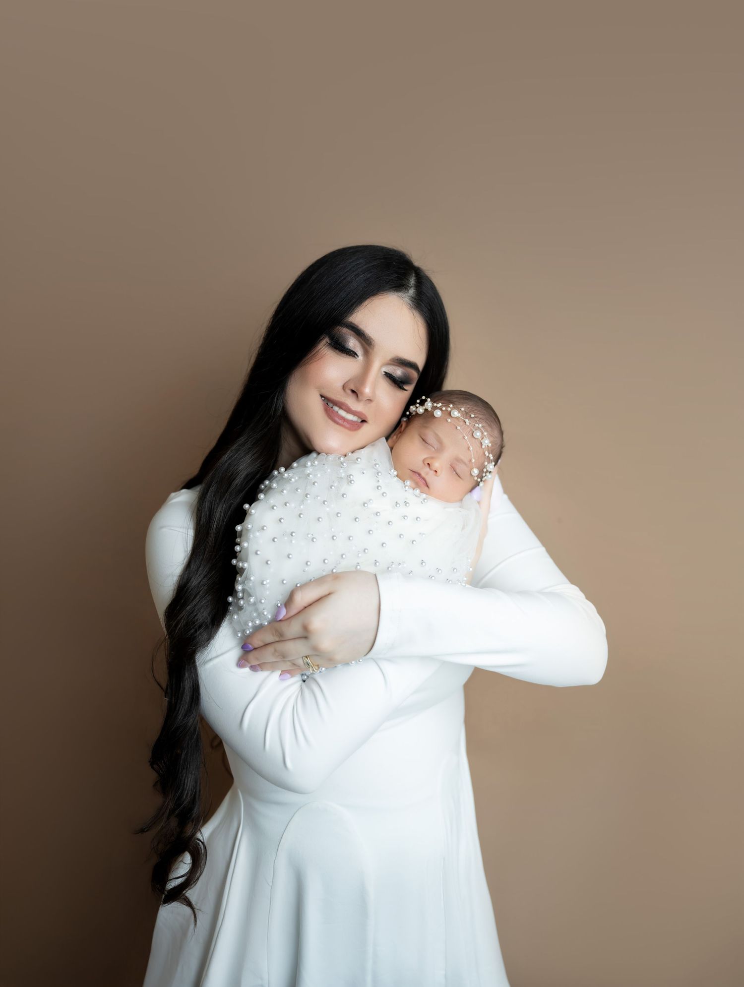A woman in a white dress holds a sleeping newborn wrapped in a pearl-adorned white swaddle against a beige background.