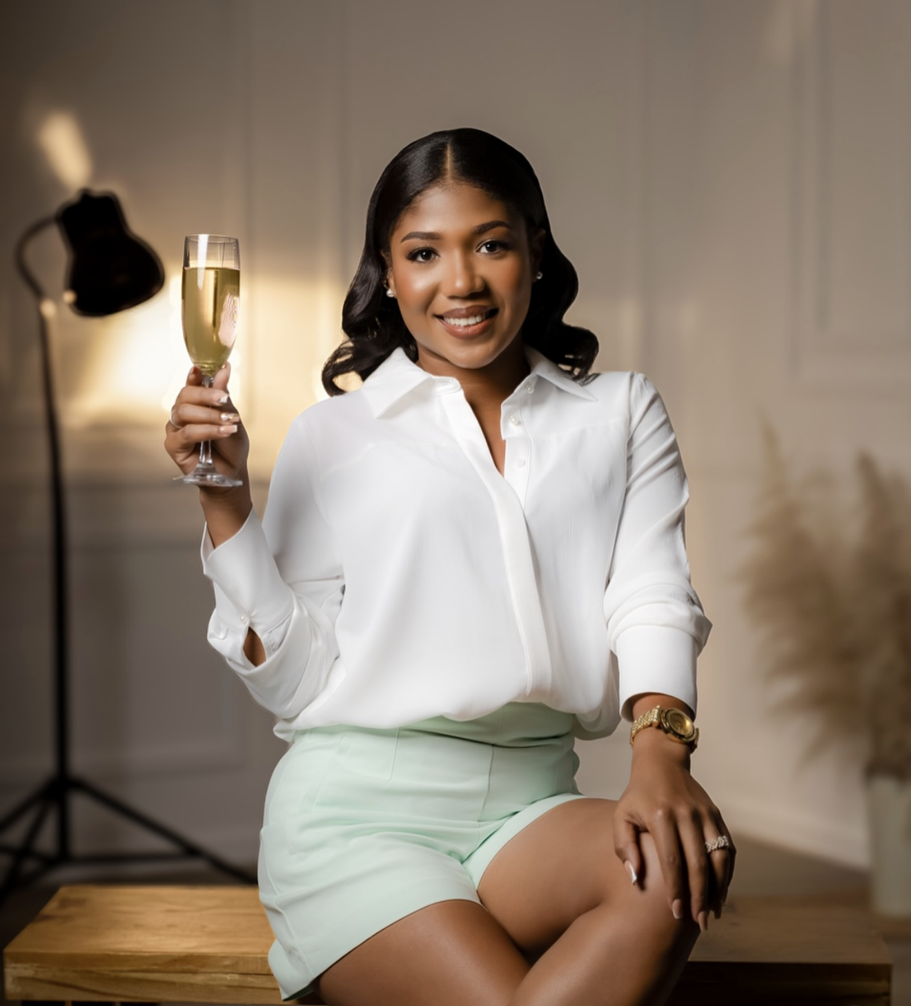 Woman in white blouse and mint skirt raising champagne glass, seated on wooden table in elegant studio setting.