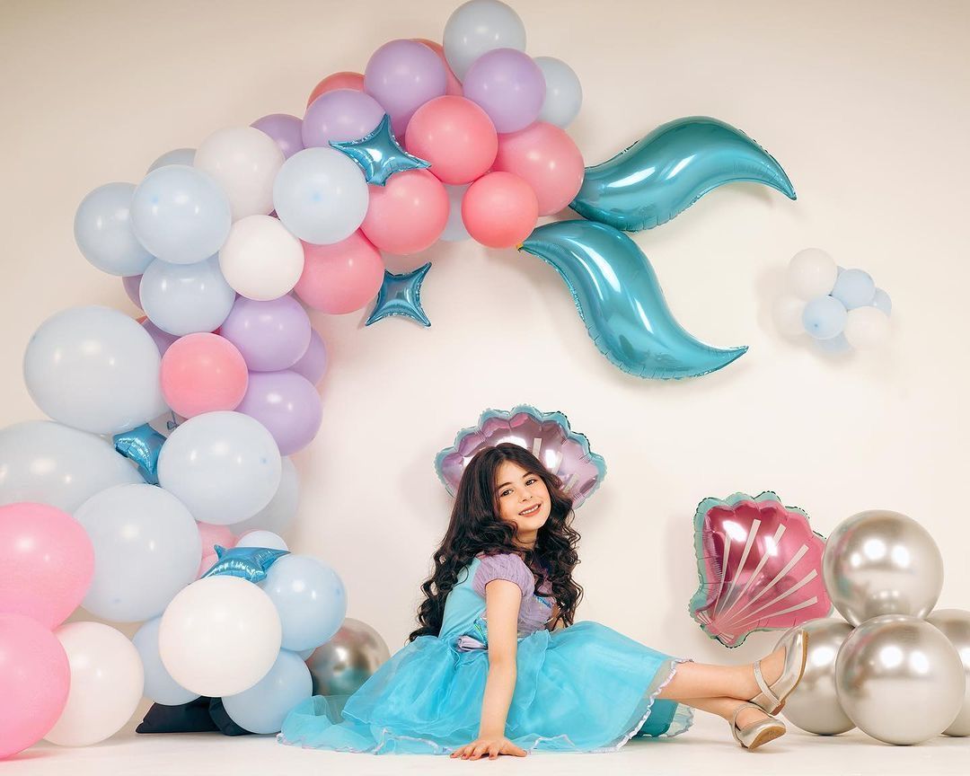 Girl in mermaid costume poses with balloon arch and mermaid tail balloon backdrop.