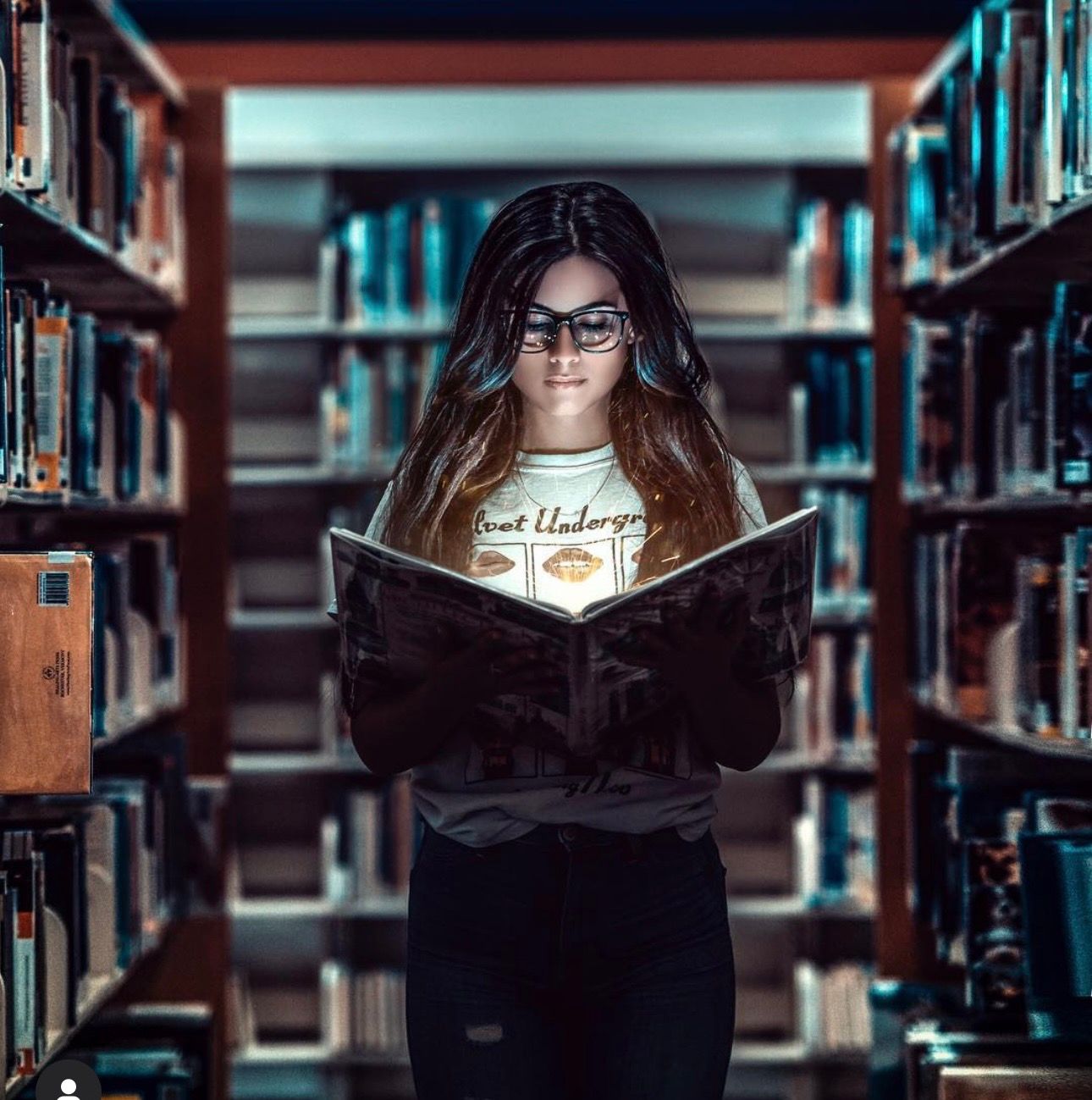A young woman with glasses reads a glowing book in a dimly lit library aisle surrounded by shelves of books.
