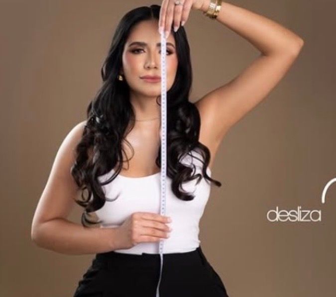 Woman with long wavy dark hair holds a measuring tape vertically from above her head to her waist, wearing a white tank top and black skirt.