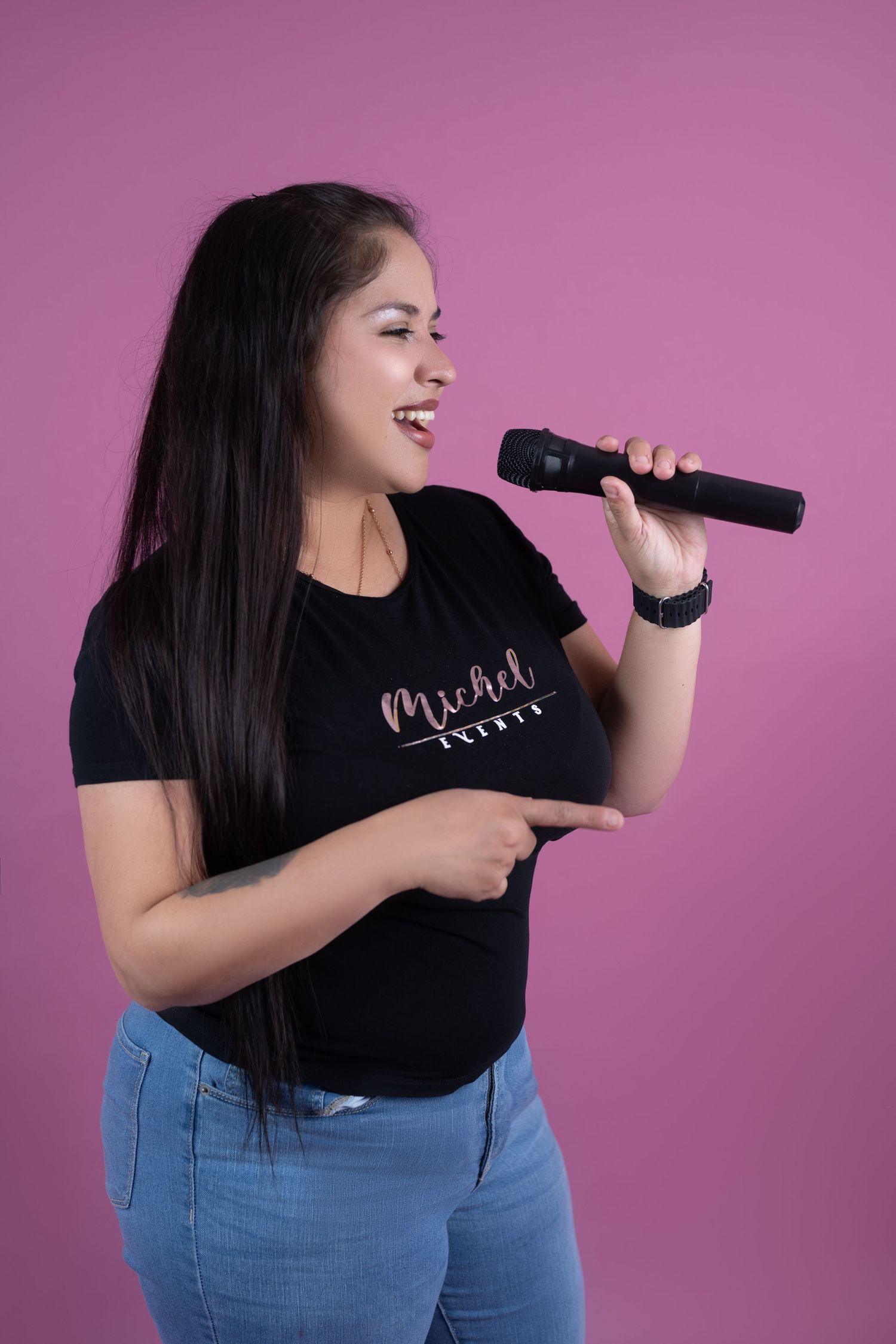 Smiling woman with long dark hair singing into a black microphone against a pink background, wearing a black graphic t-shirt and jeans.