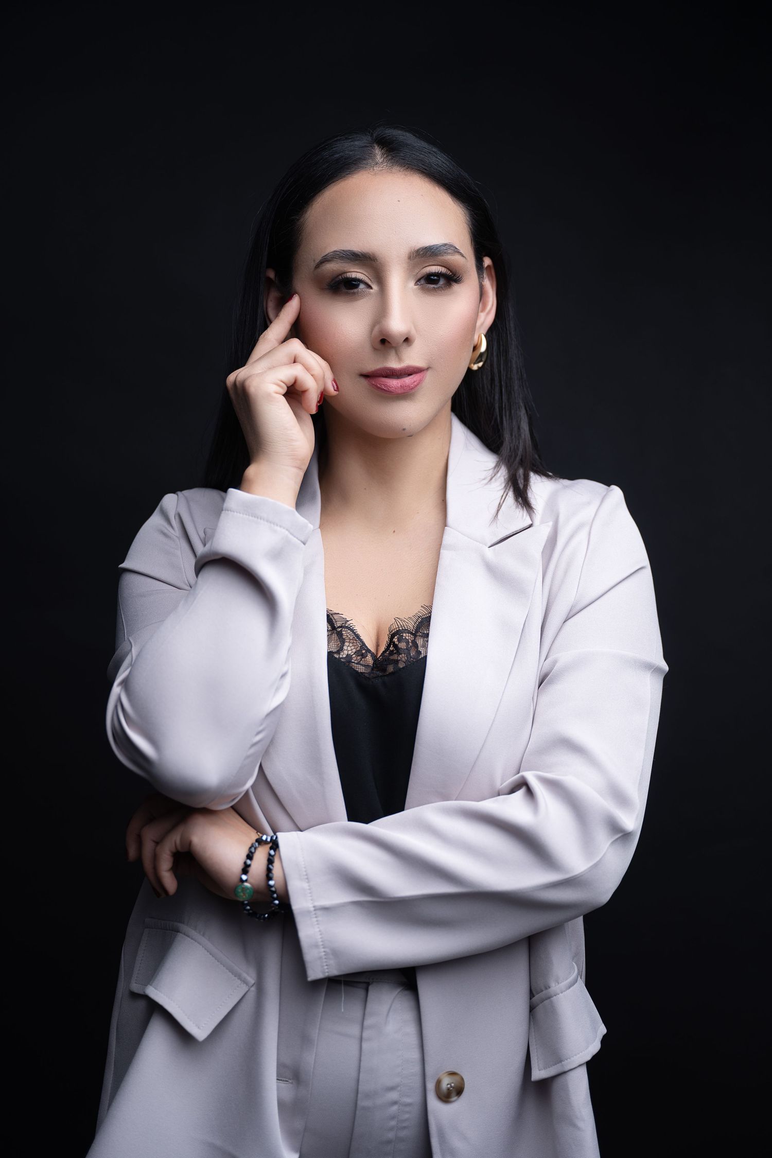 Woman with long dark hair in a light gray blazer and black lace top poses with her hand at her temple against a black background.