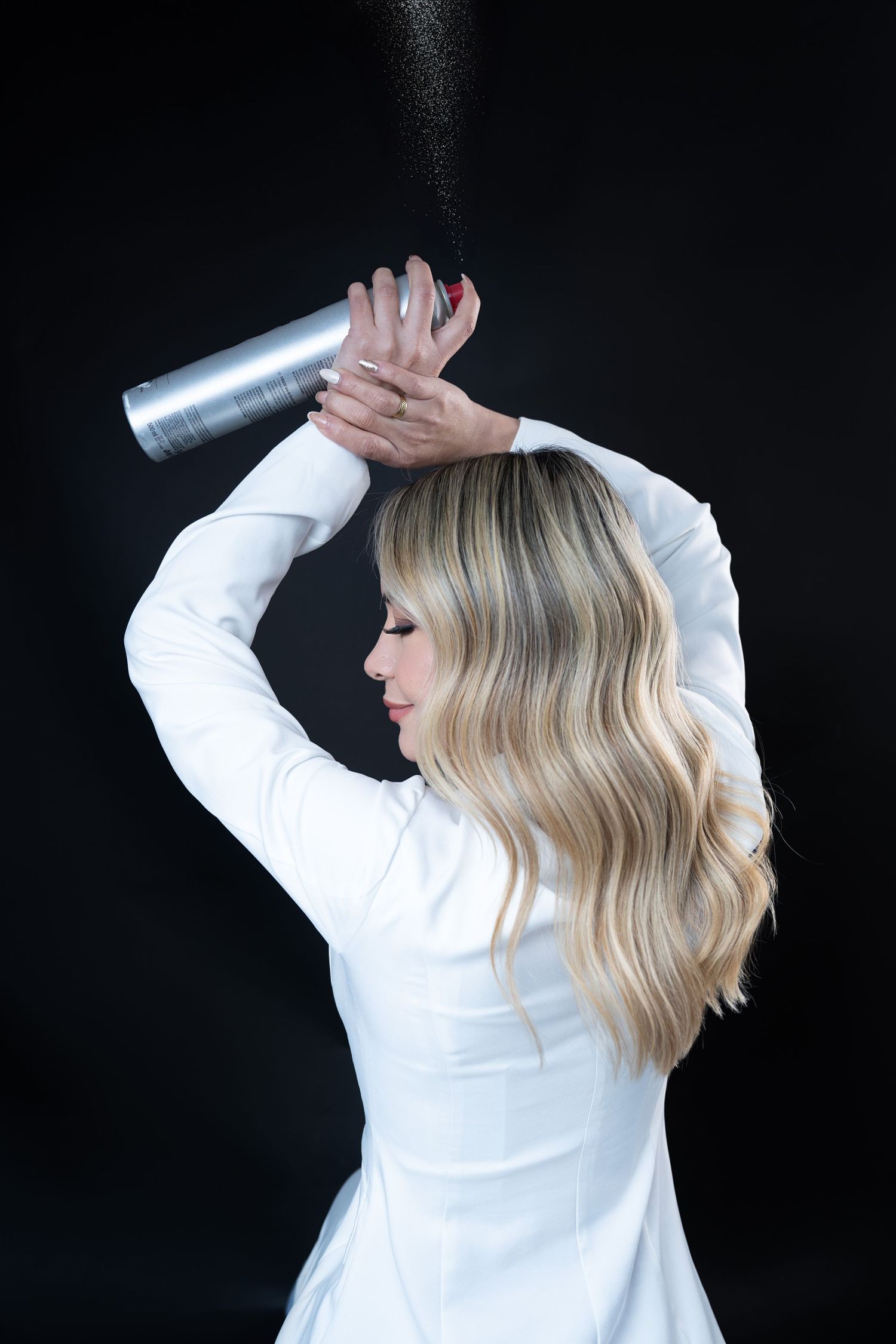 Blonde woman in white dress applying hairspray overhead against black background, showcasing wavy hair styling technique.