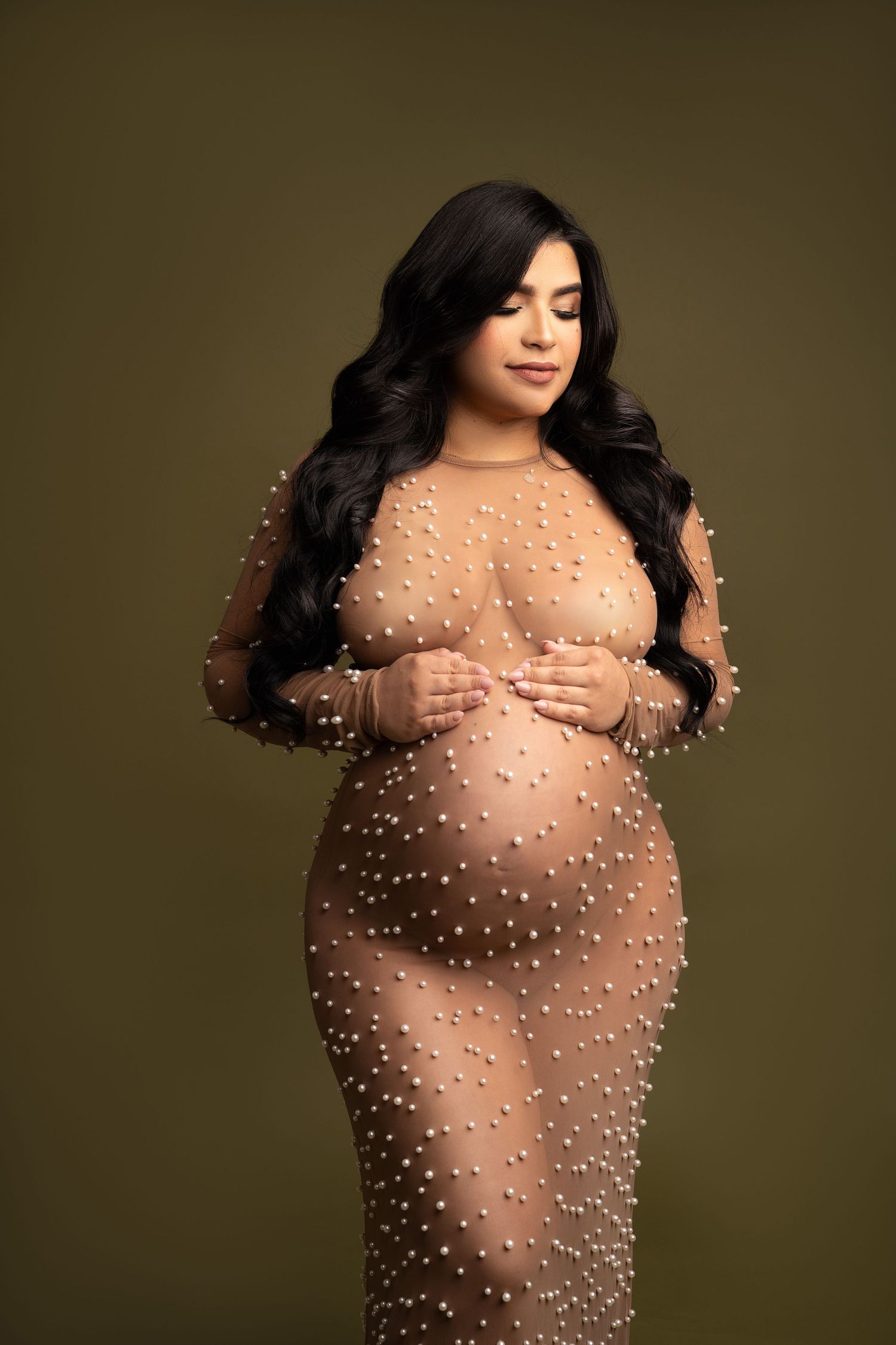 Maternity portrait featuring sheer pearl-adorned gown against dark background.