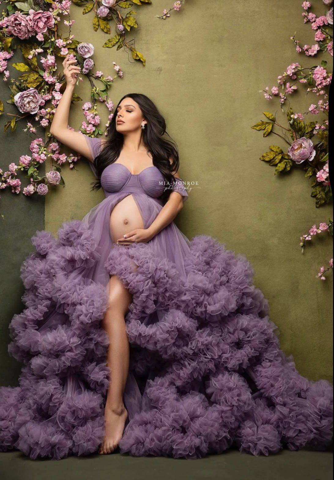 Pregnant woman in lavender ruffle tulle maternity gown posing with pink flowers against green backdrop.