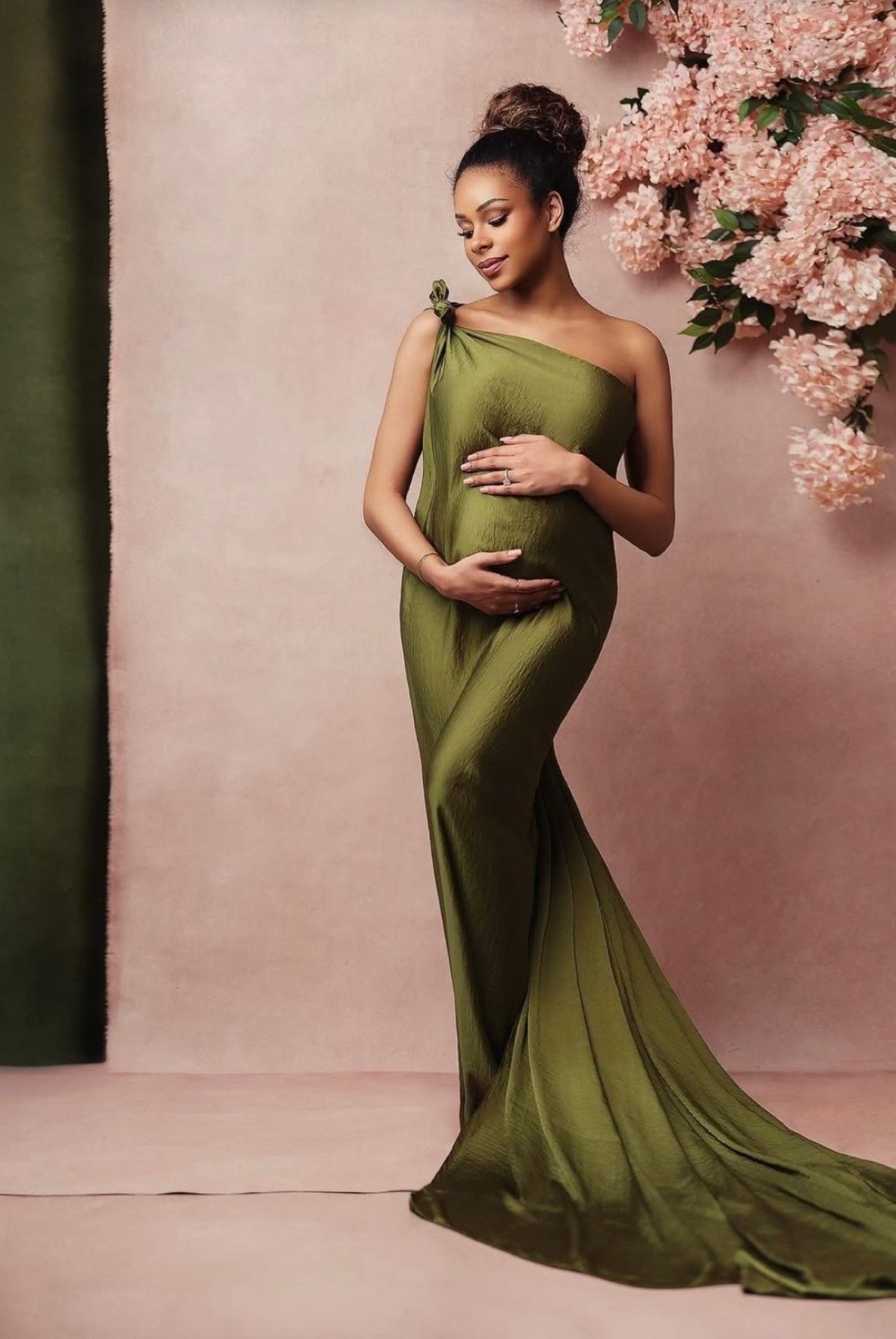 Pregnant woman in elegant olive green one-shoulder maternity gown posing gracefully against a soft pink floral backdrop.