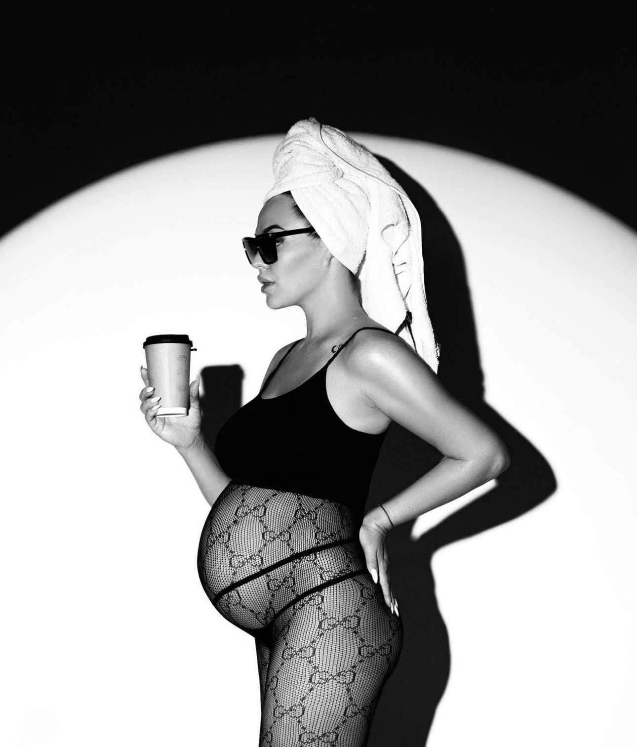 Black and white photo of a pregnant woman in sunglasses, towel turban, holding coffee cup, wearing Gucci tights.