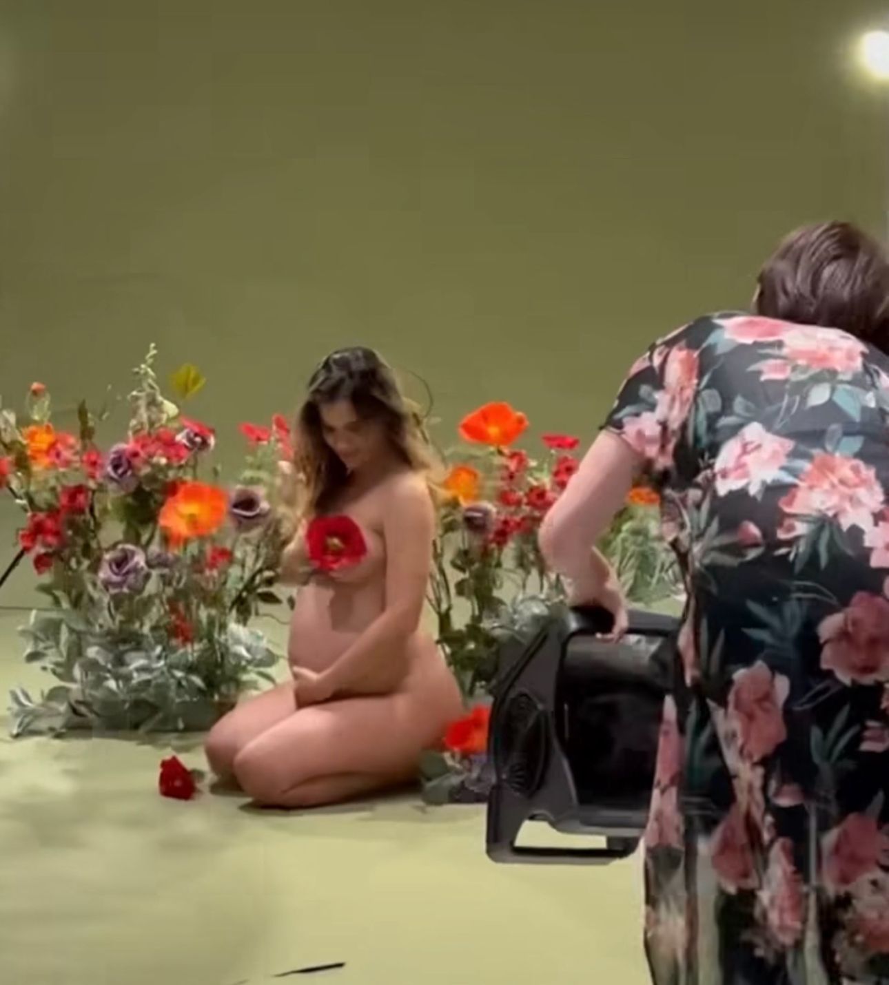 Pregnant woman posing nude among colorful flowers on green backdrop during professional photo shoot.