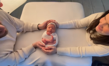 A newborn baby wearing a striped outfit lies on a white couch while being cradled.