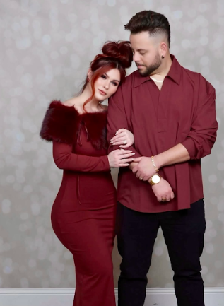 A couple in matching burgundy outfits pose together against a gray background for a romantic portrait.
