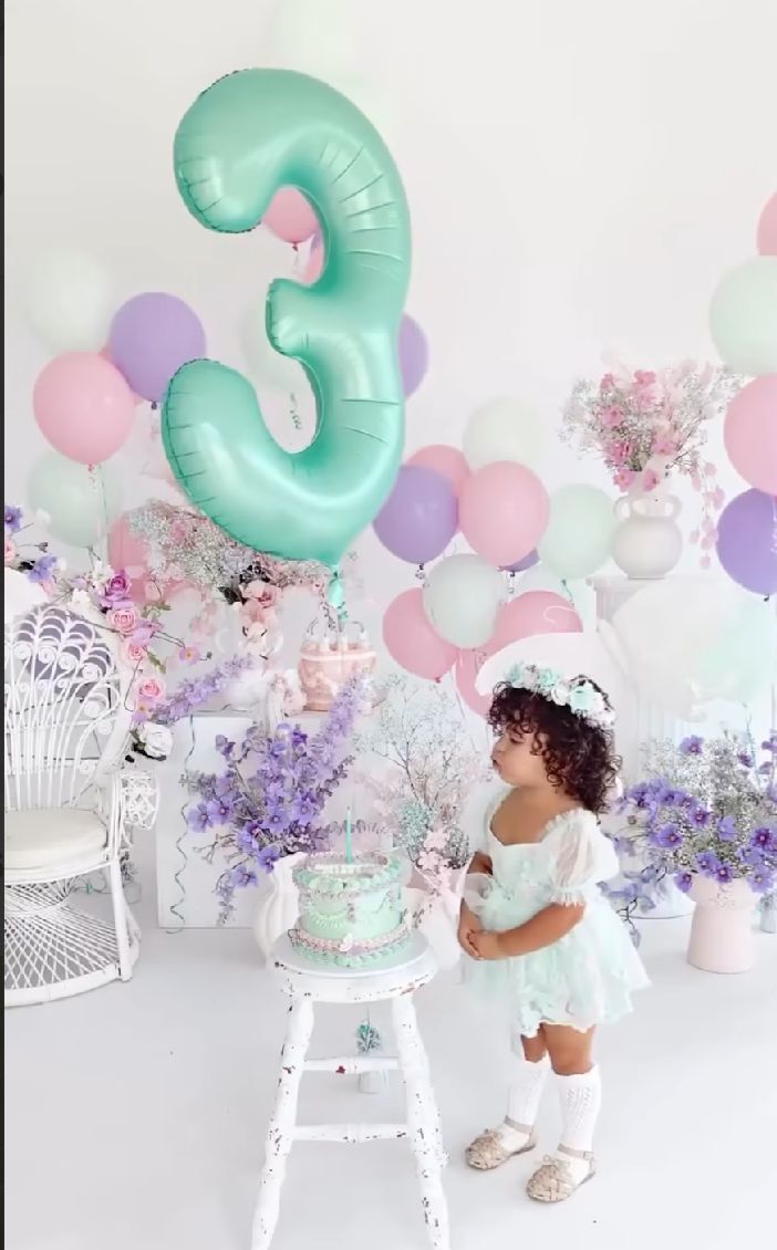 Birthday party setup with mint green number 3 balloon, pastel decorations, purple flowers and white furniture for toddler celebration.