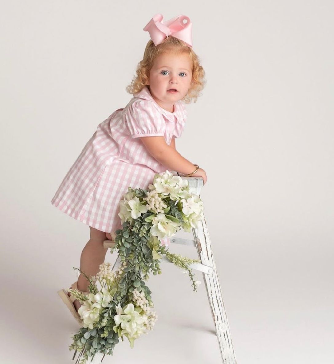 Sweet photo series showing a toddler in pink gingham dress playing with white flowers on a white stool.