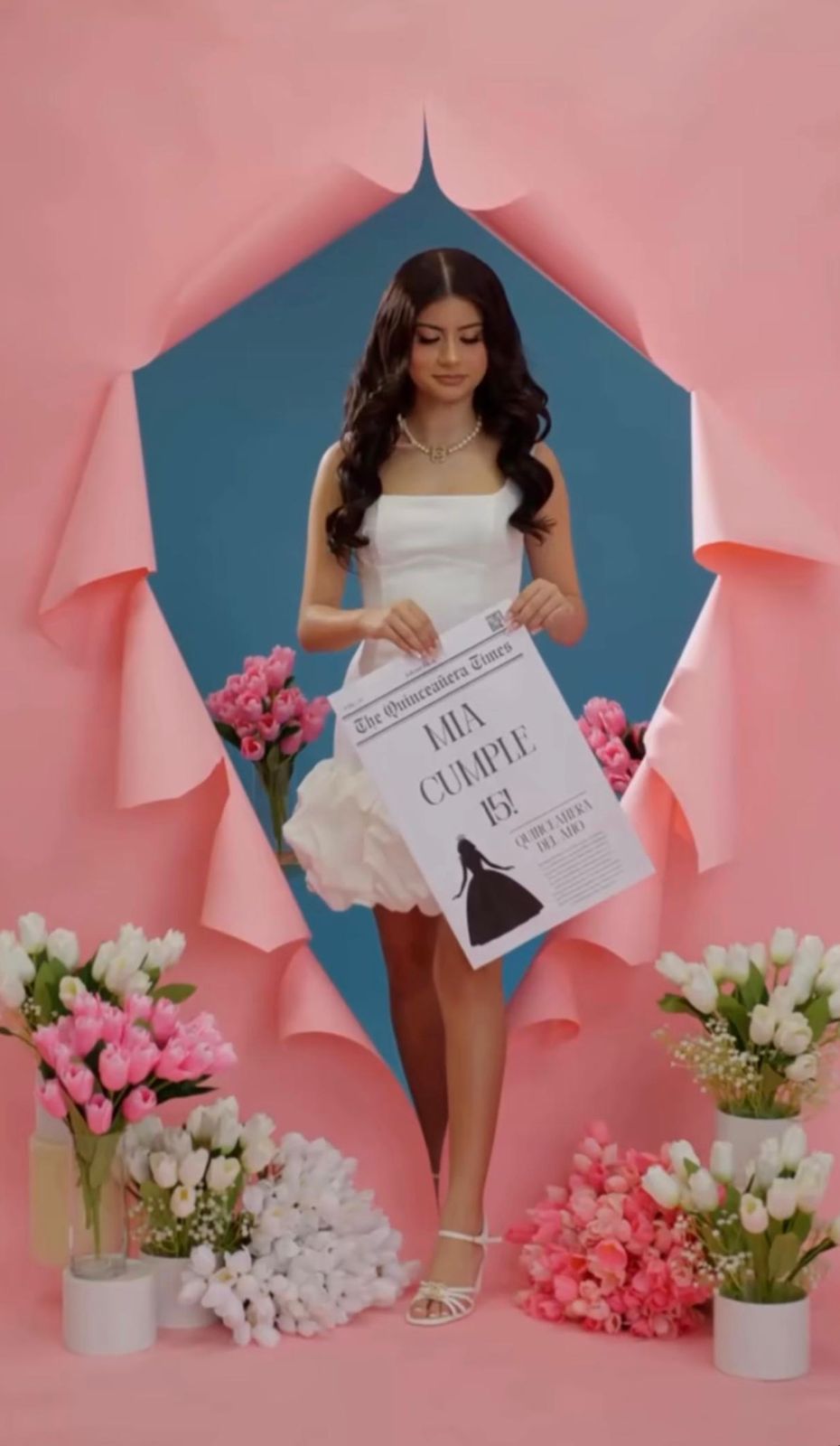 Young woman in white dress holds a quinceañera newspaper prop, surrounded by pink tulips against a pink and blue backdrop.