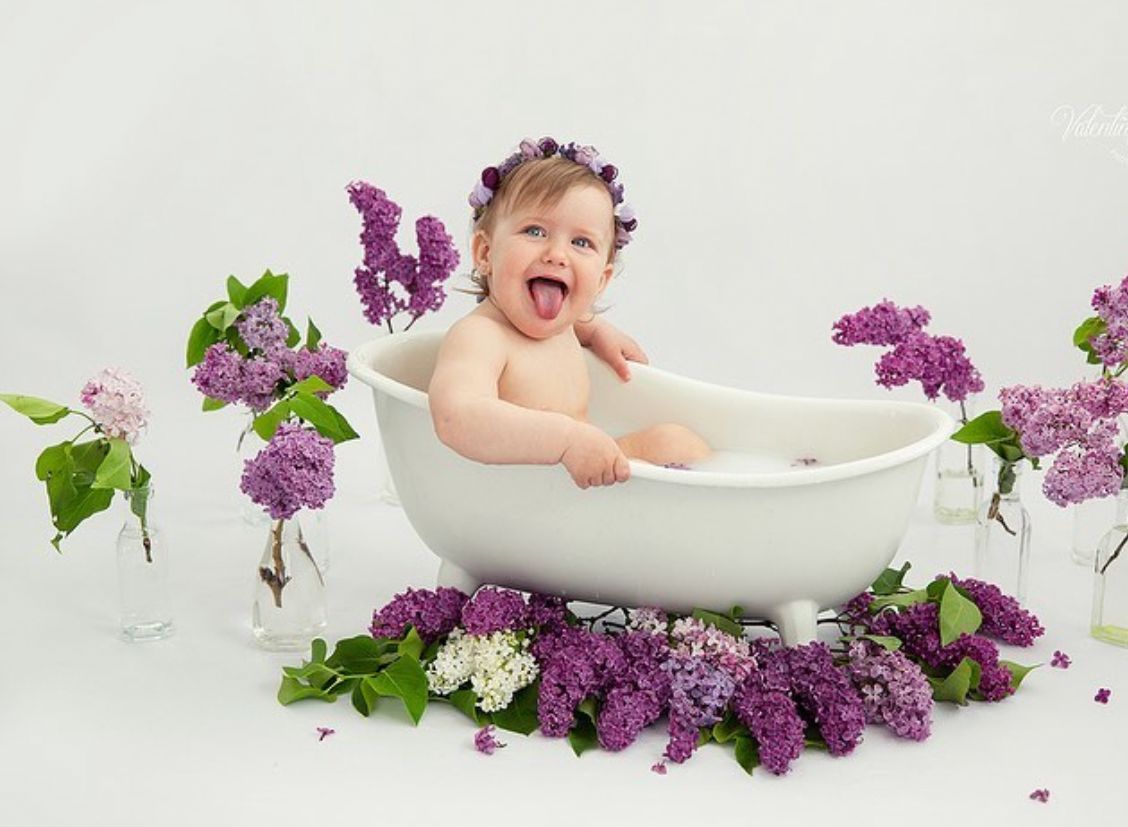 A joyful baby sits in a white bathtub surrounded by vibrant purple and white lilac flowers.