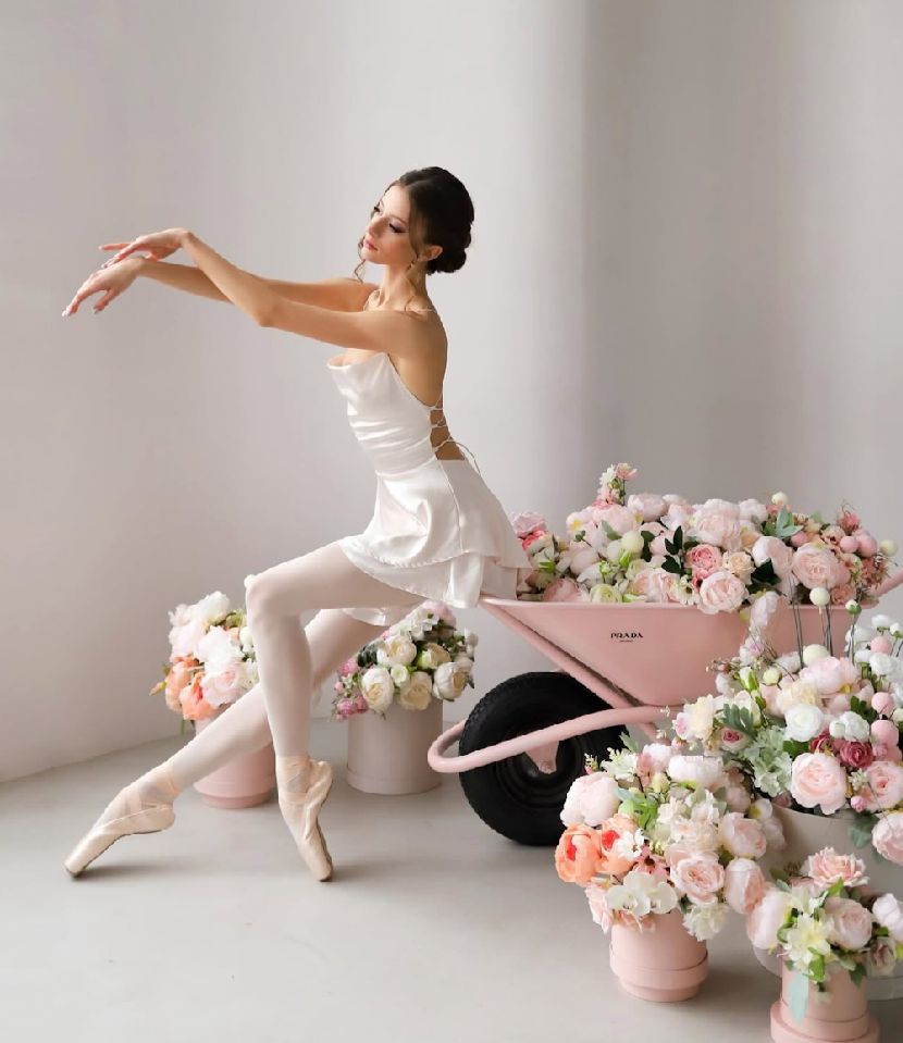Ballet dancer in white tutu poses gracefully among pink and white floral arrangements.