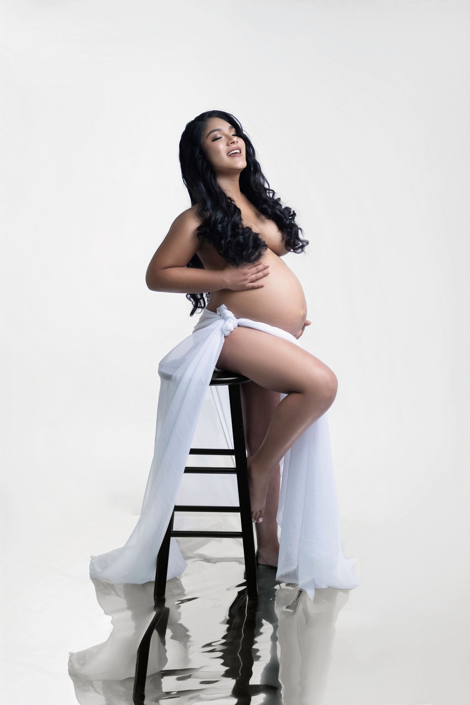 Pregnant woman posing gracefully in white draped fabric on a stool against a white studio background.