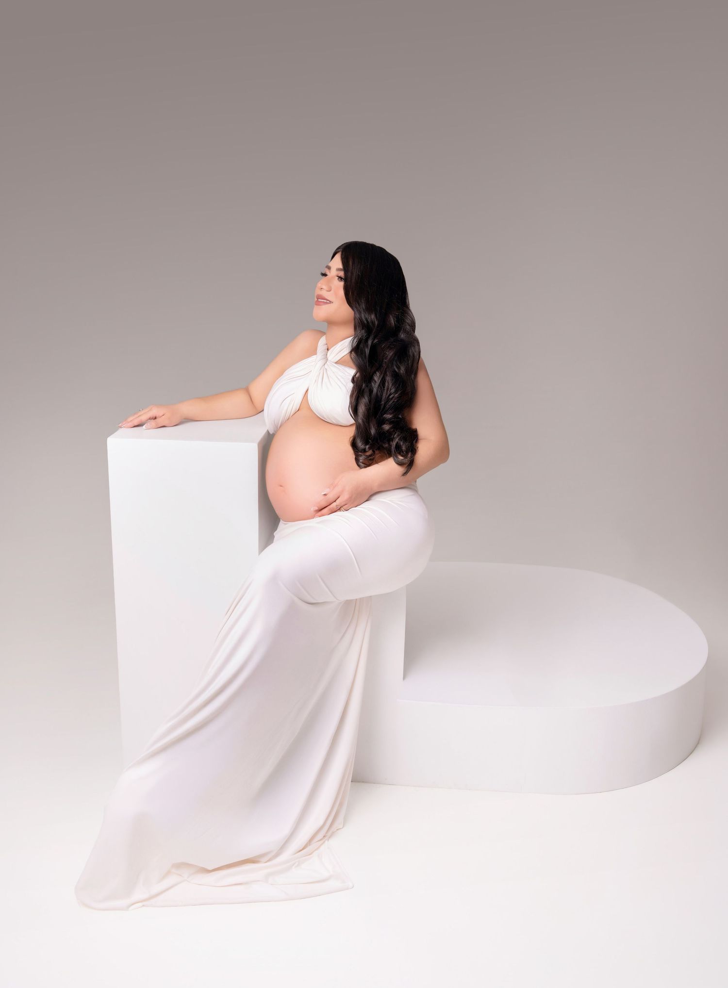 Pregnant woman in white halter crop top and skirt poses elegantly against white geometric props in a studio.
