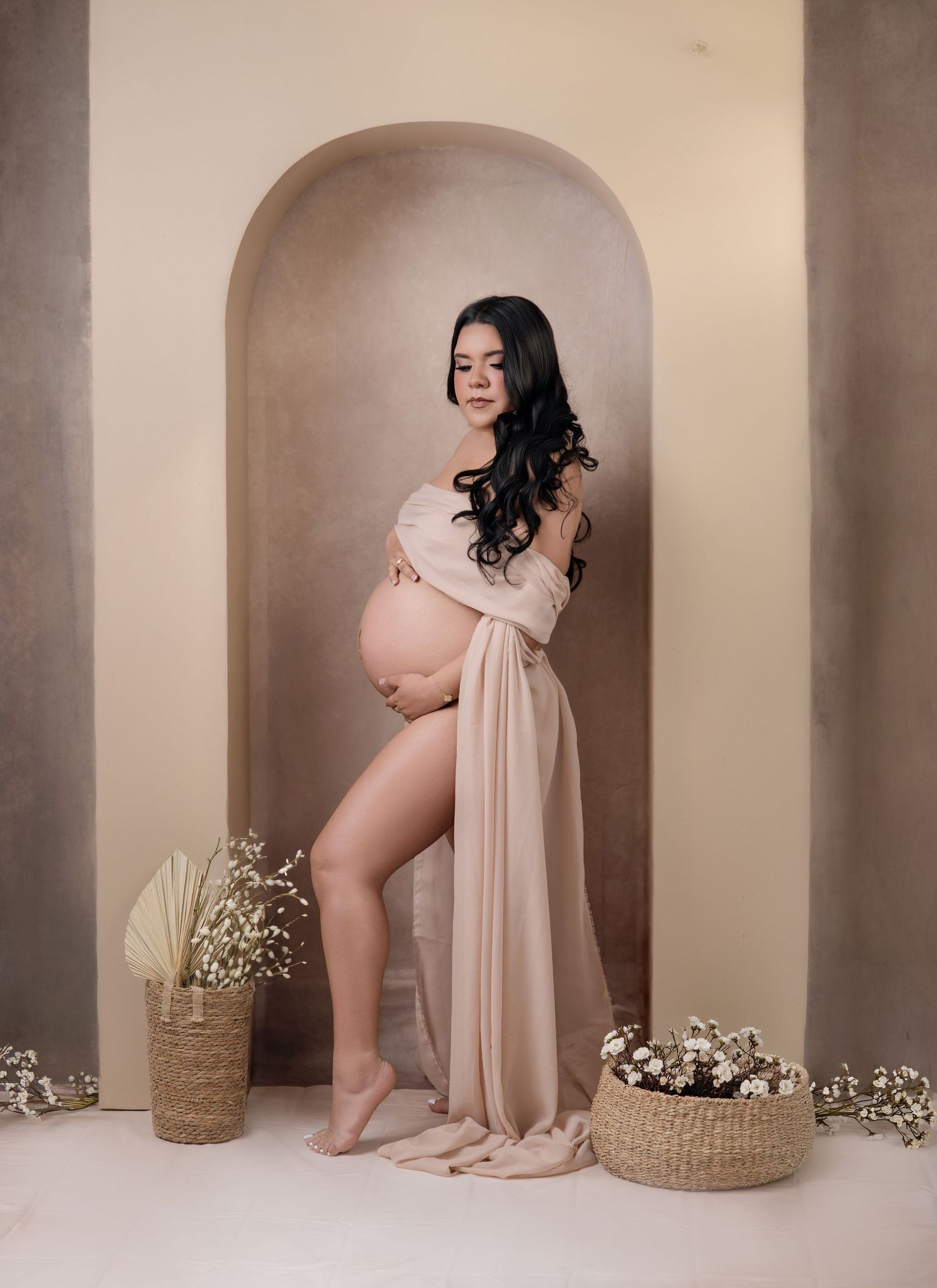 Pregnant woman in blush draped fabric poses barefoot in arched alcove with floral baskets in a maternity photo shoot.