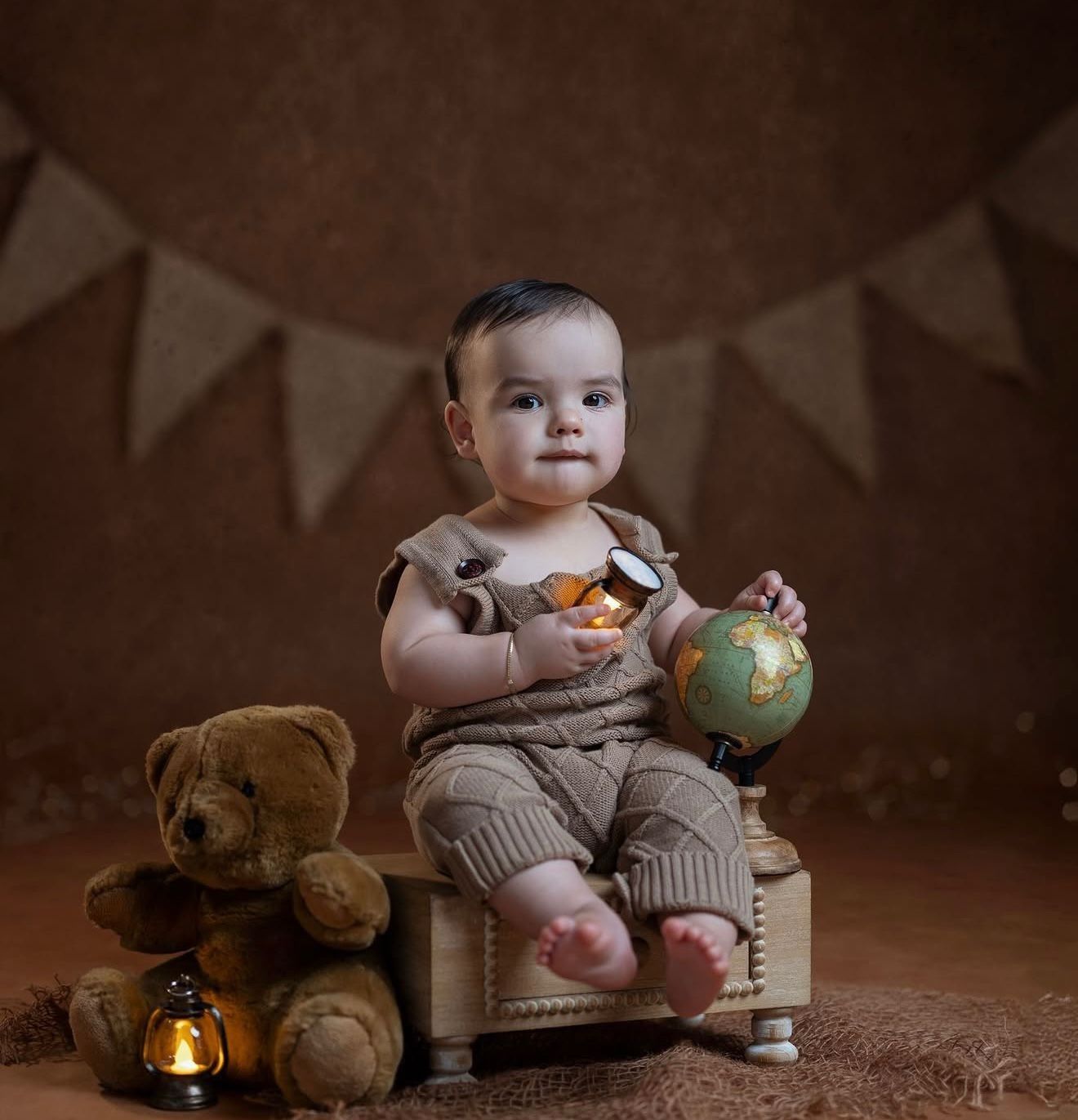 Adorable baby in brown outfit sits on small wooden box holding a globe, with teddy bear and lantern nearby.