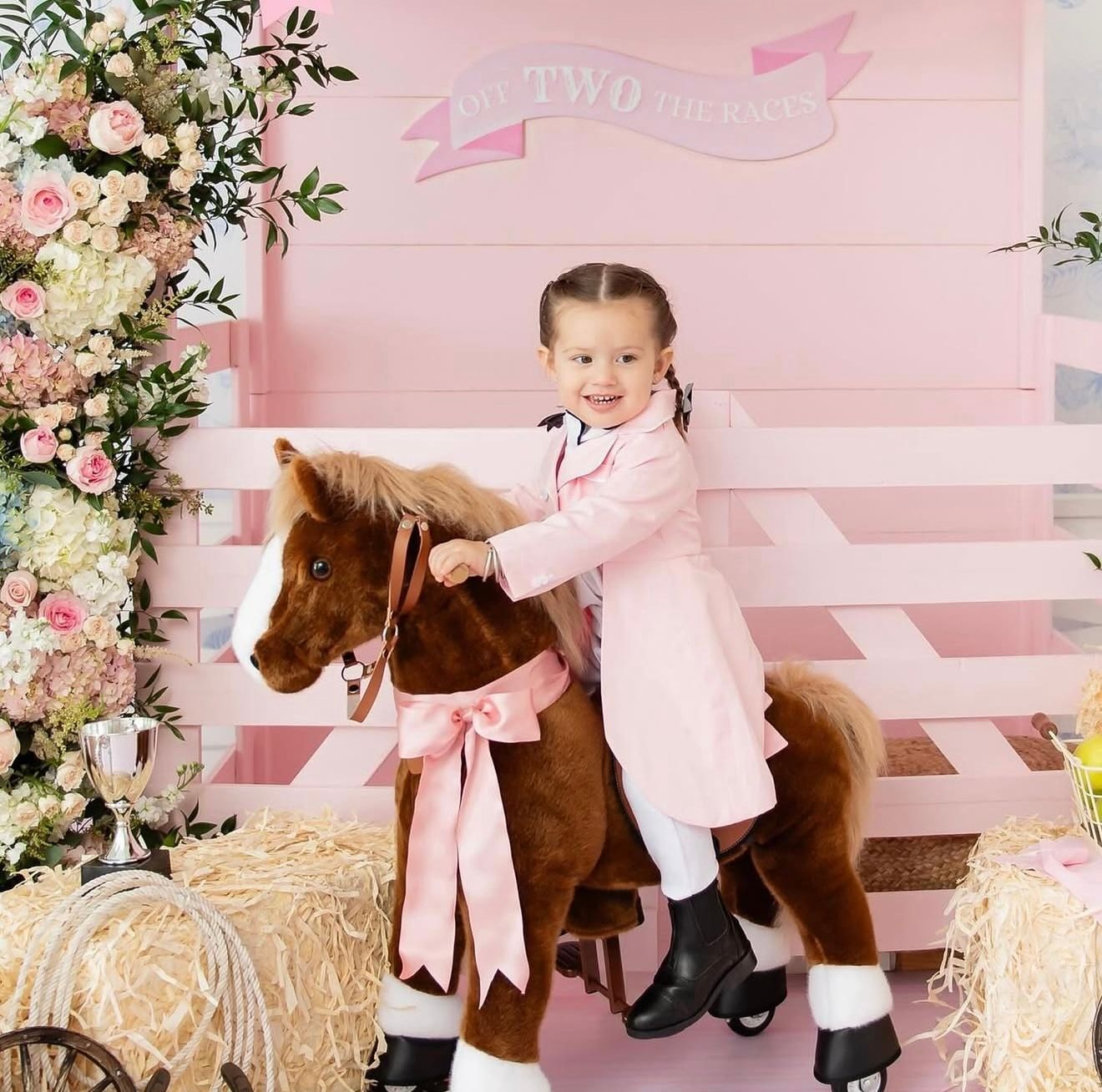 A young girl in pink coat and boots sits on a decorative toy horse at a pink-themed second birthday party.