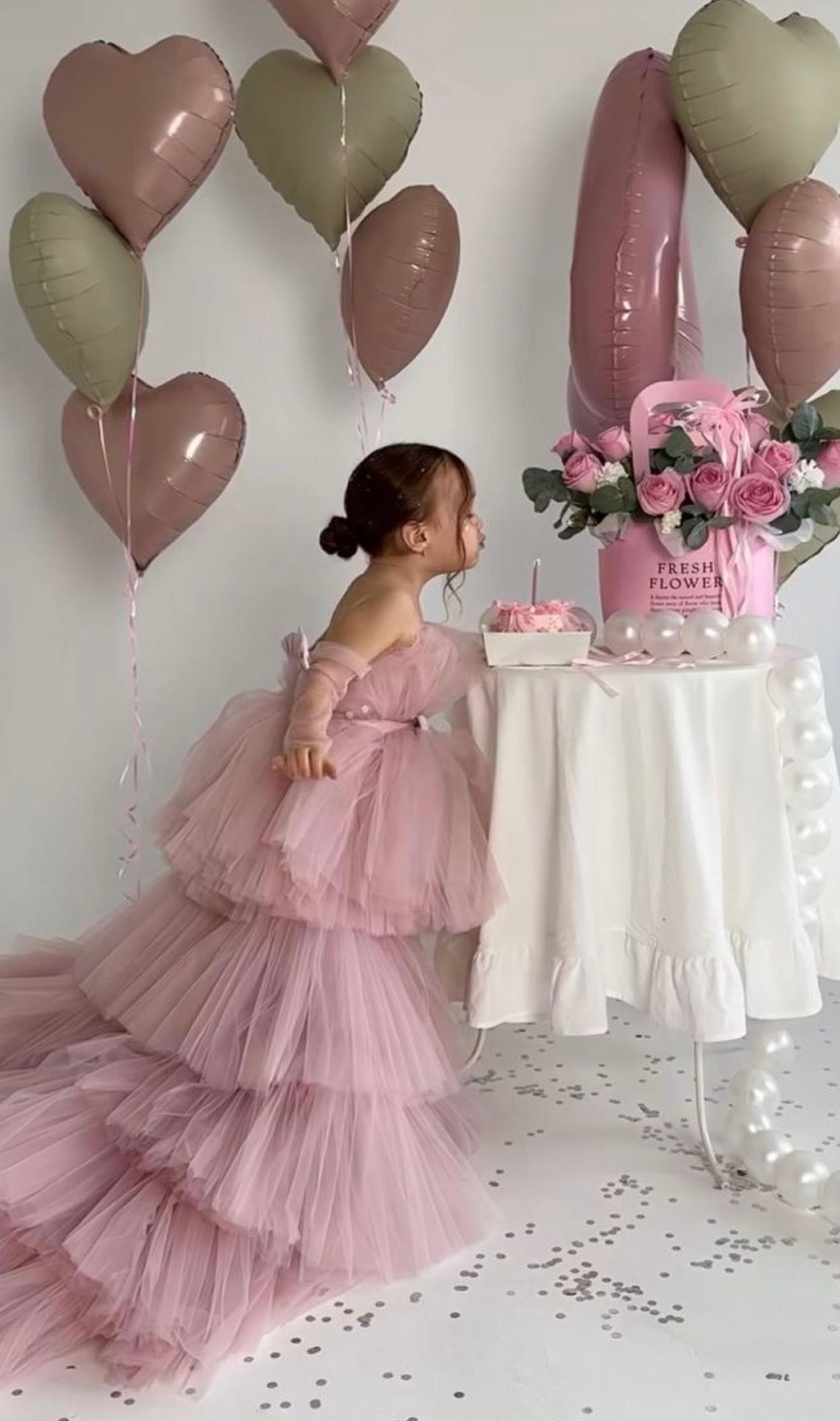 A little girl in a pink tulle dress stands by a birthday table with heart balloons and fresh flowers.