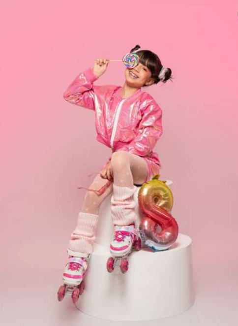 Girl in pink shiny jacket and roller skates sits on white pedestal holding lollipop near number 8 balloon.