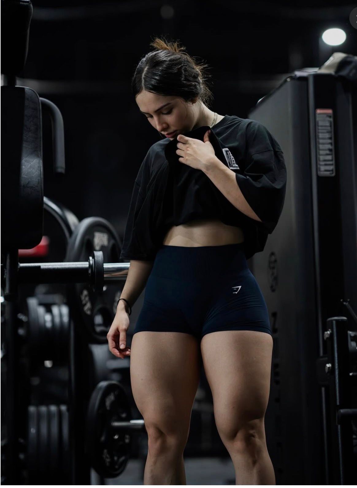 Woman in black crop top and Gymshark shorts standing in dark gym near barbell rack.