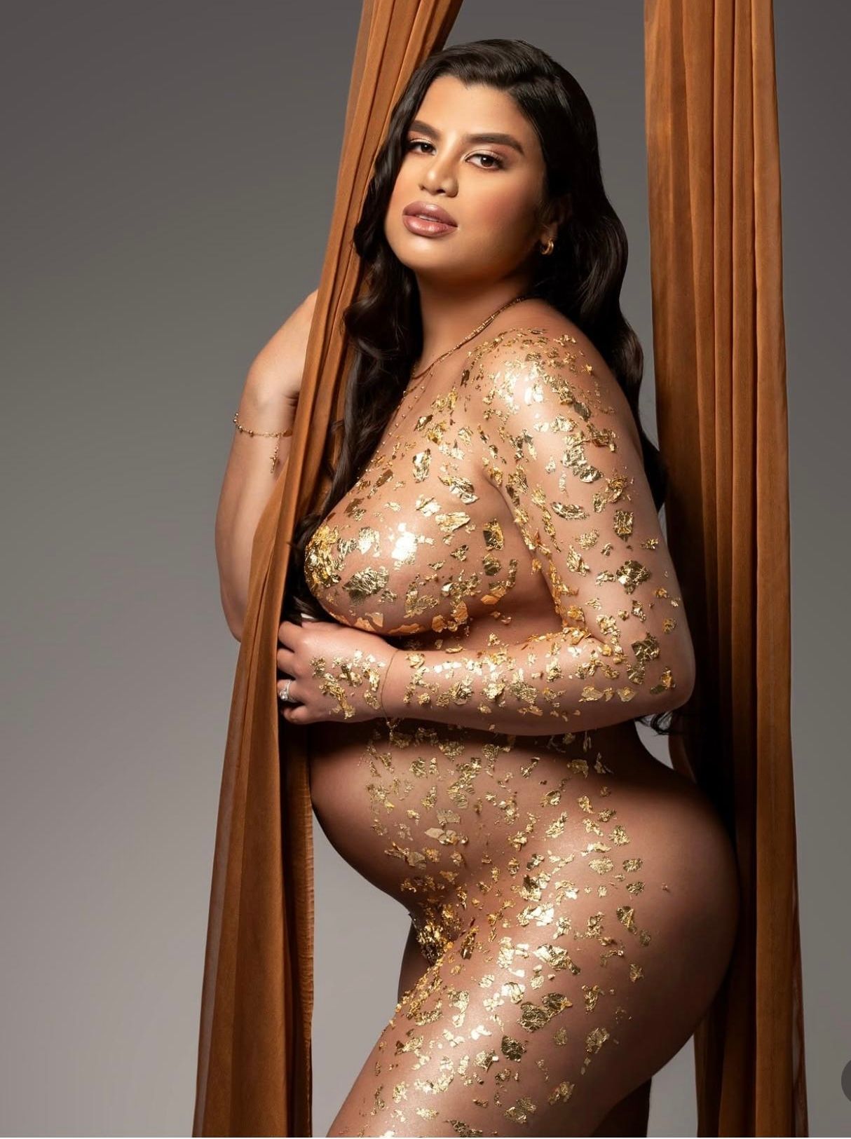 Pregnant woman in gold glitter body paint posing elegantly with flowing orange fabric backdrop.